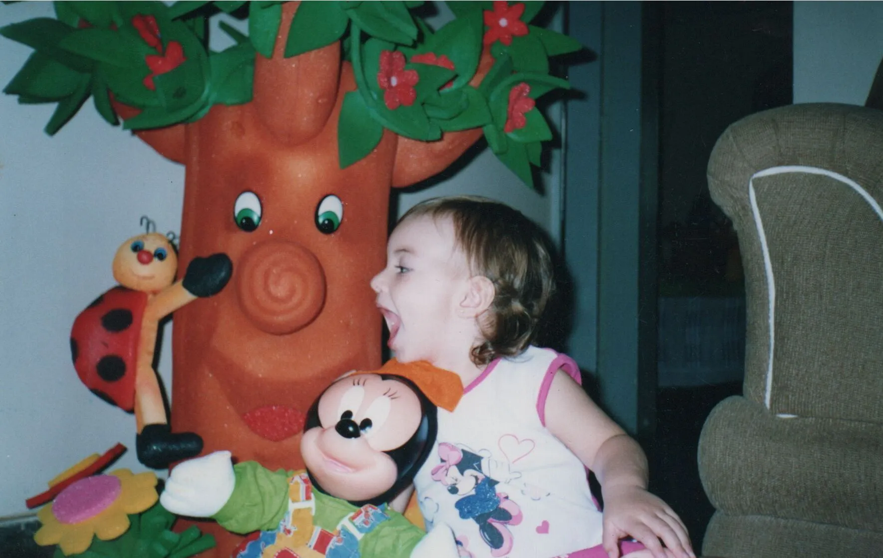 Uma criança sorridente com uma camisa da Minnie Mouse senta-se ao lado de um suporte de árvore de desenho animado e um brinquedo de pelúcia da Minnie, com a boca aberta de admiração.