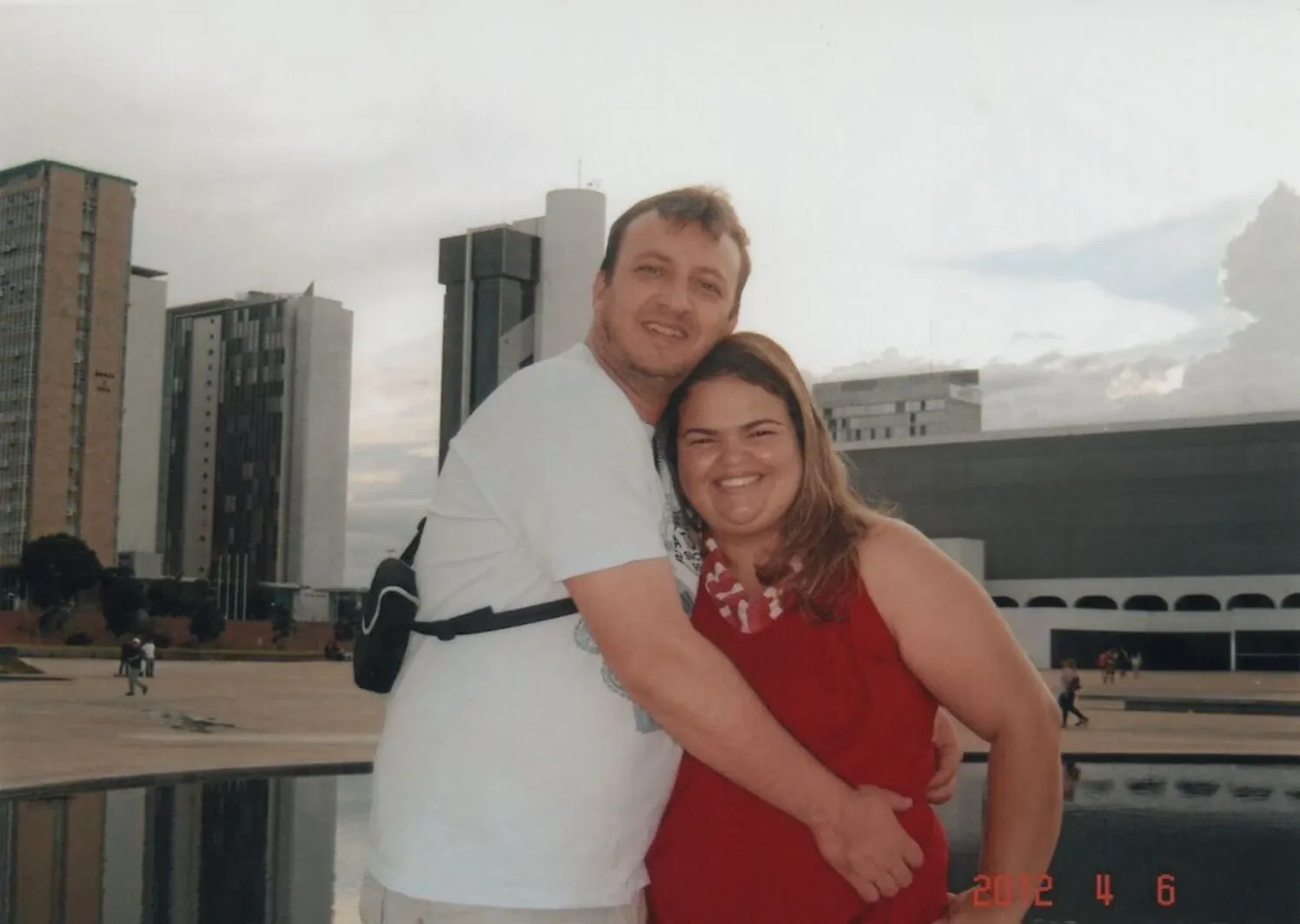 Um homem e uma mulher abraçados e sorrindo para a câmera, com prédios de arquitetura moderna e um céu parcialmente nublado ao fundo.