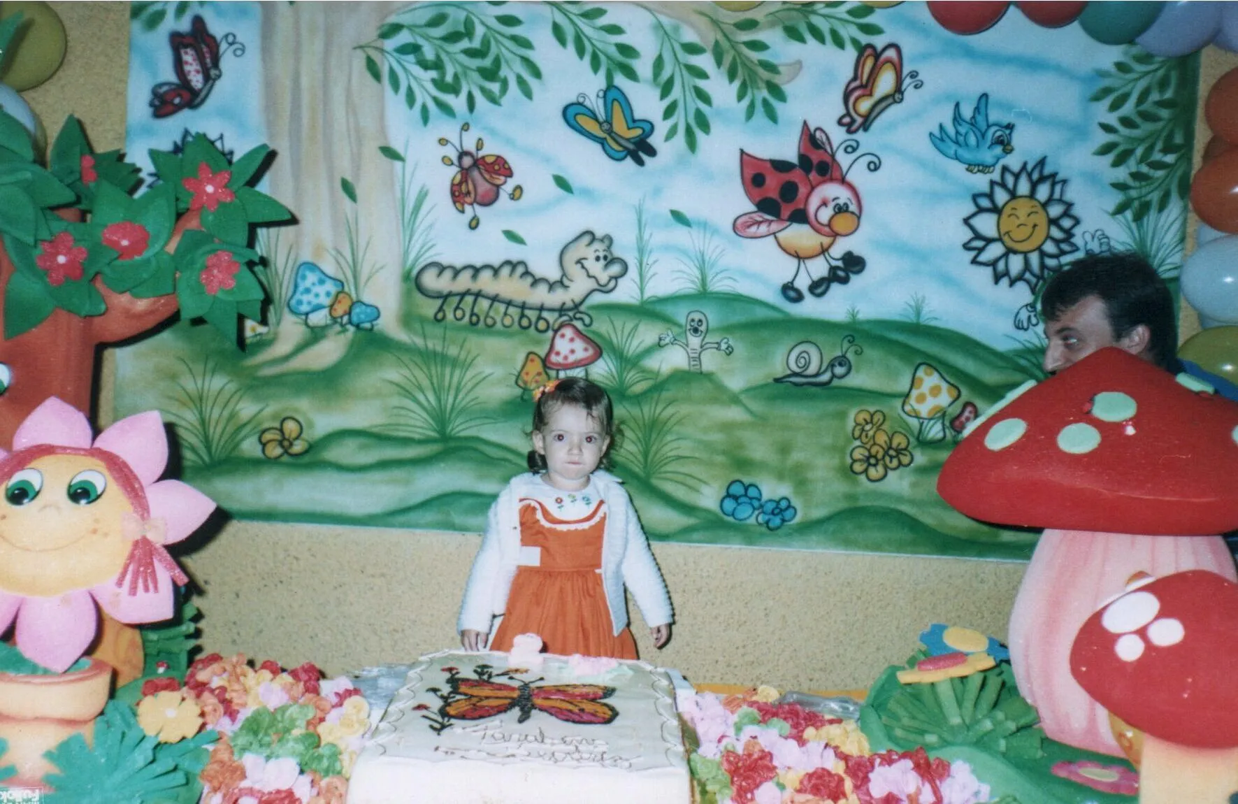 Menina pequena de vestido laranja e casaquinho branco parada na frente de um bolo de aniversário branco decorado com uma borboleta, com um mural de jardim de desenho animado e decoração de festa ao fundo.