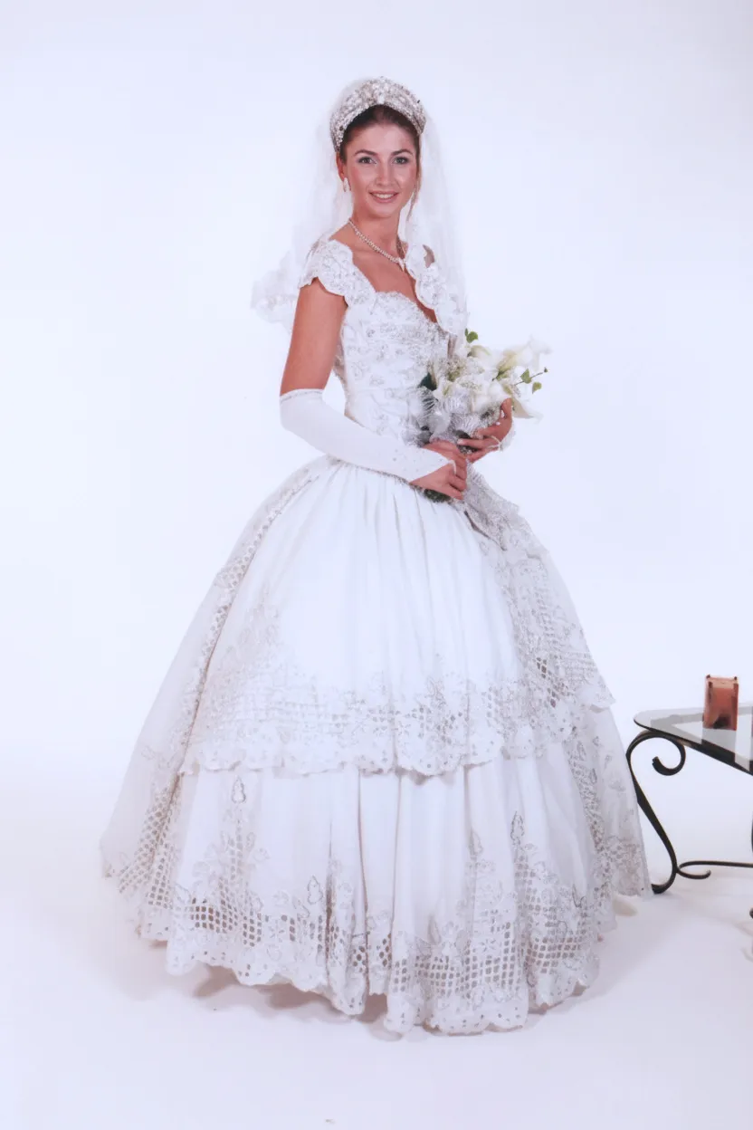 Noiva sorridente em um vestido de noiva branco tradicional, com véu e luvas brancas, segurando um buquê de flores brancas.