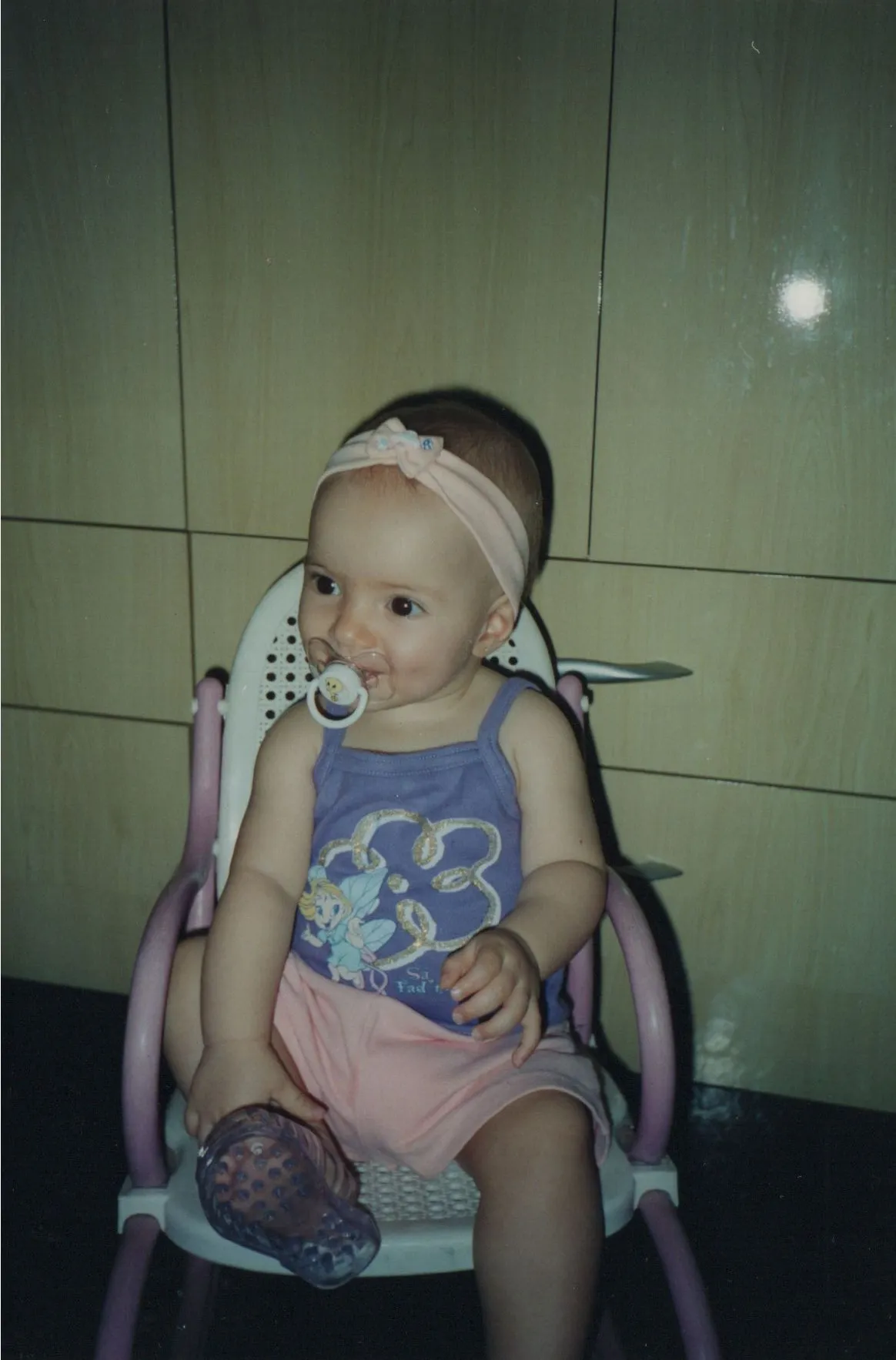 Bebê caucasiano com laço rosa na cabeça, babador e uma blusa roxa, sentado em uma cadeira de alimentação. (Original)