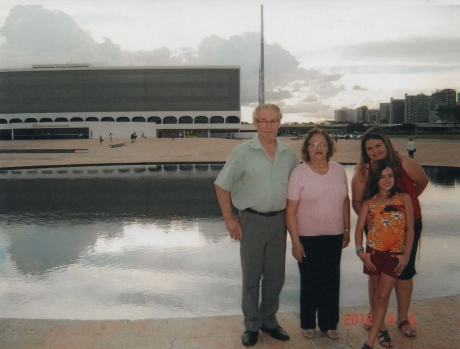 Um grupo familiar de quatro pessoas, um homem e três mulheres de diferentes gerações, posa à beira de um corpo d'água refletindo nuvens. Edifícios modernos e um céu parcialmente nublado formam o pano de fundo.