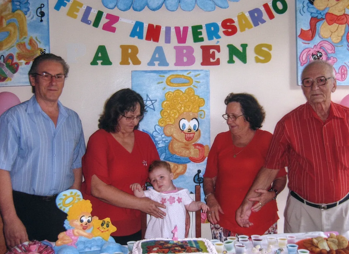 Um grupo de cinco adultos e uma criança, todos vestindo roupas brilhantes, posa alegremente em torno de um bolo de aniversário branco decorado com uma flor vermelha, em uma sala decorada com um mural de "FELIZ ANIVERSÁRIO, PARABÉNS" em várias cores e balões rosa e azul.