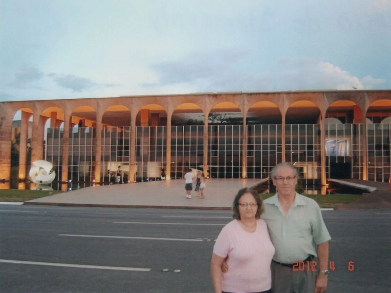 Dois idosos posam em frente a um grande edifício com arcadas e janelas reflexivas em um dia nublado.