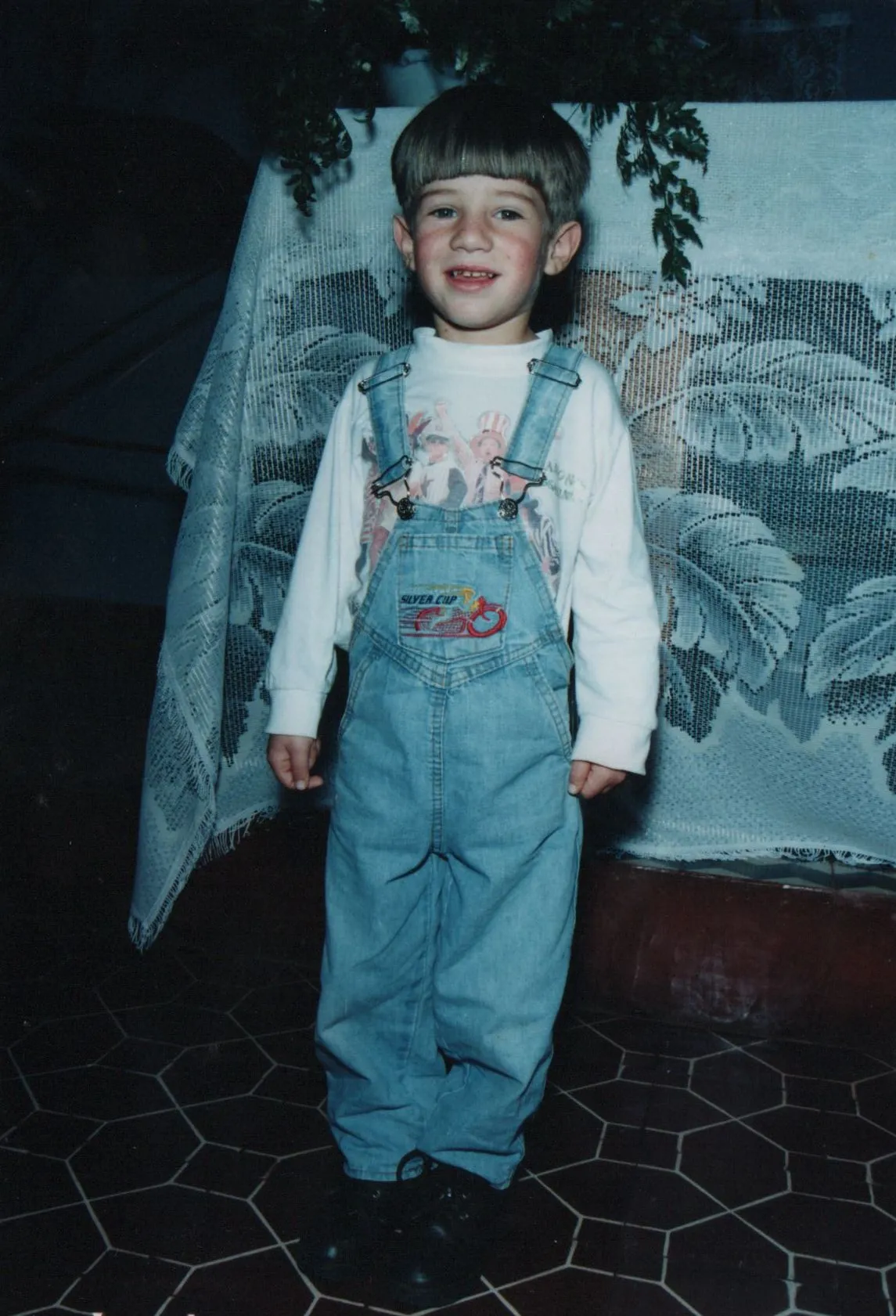 Criança pequena branca sorrindo e de pé, usando jardineira jeans sobre uma camiseta branca, na frente de uma mesa coberta com uma toalha de renda branca e folhagem verde escura. (Original)