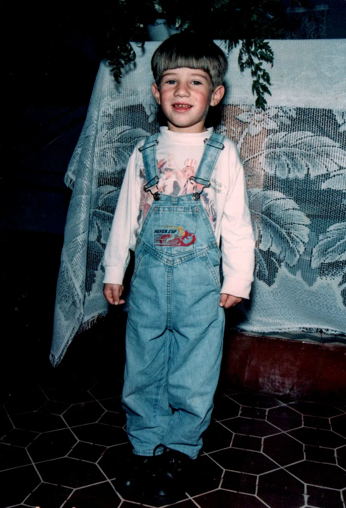 Criança pequena branca sorrindo e de pé, usando jardineira jeans sobre uma camiseta branca, na frente de uma mesa coberta com uma toalha de renda branca e folhagem verde escura.