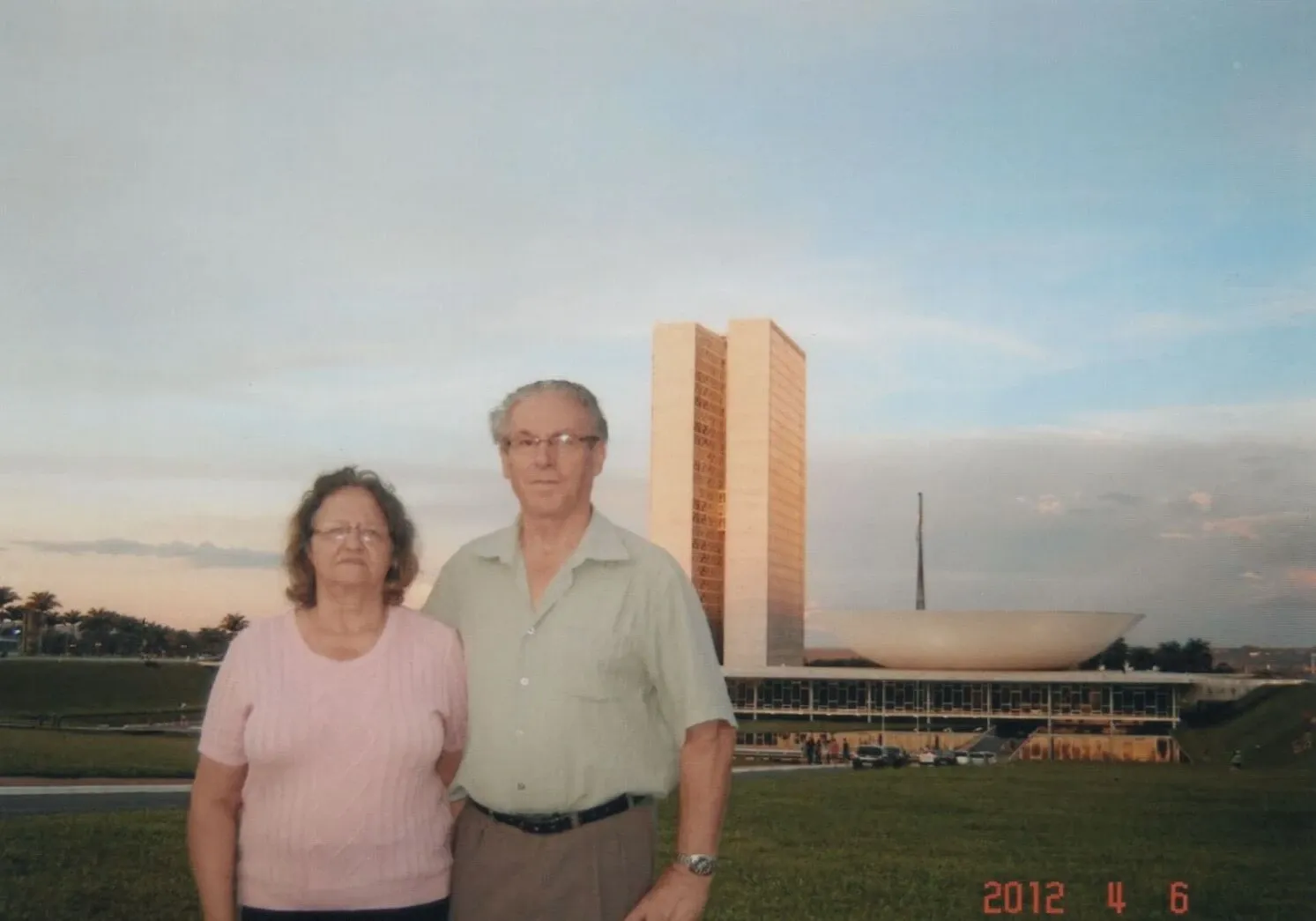 Um casal mais velho está na frente do Congresso Nacional em Brasília, sob um céu claro e crepuscular, com um homem de óculos e camisa verde-clara ao lado de uma mulher com óculos e camisa rosa.