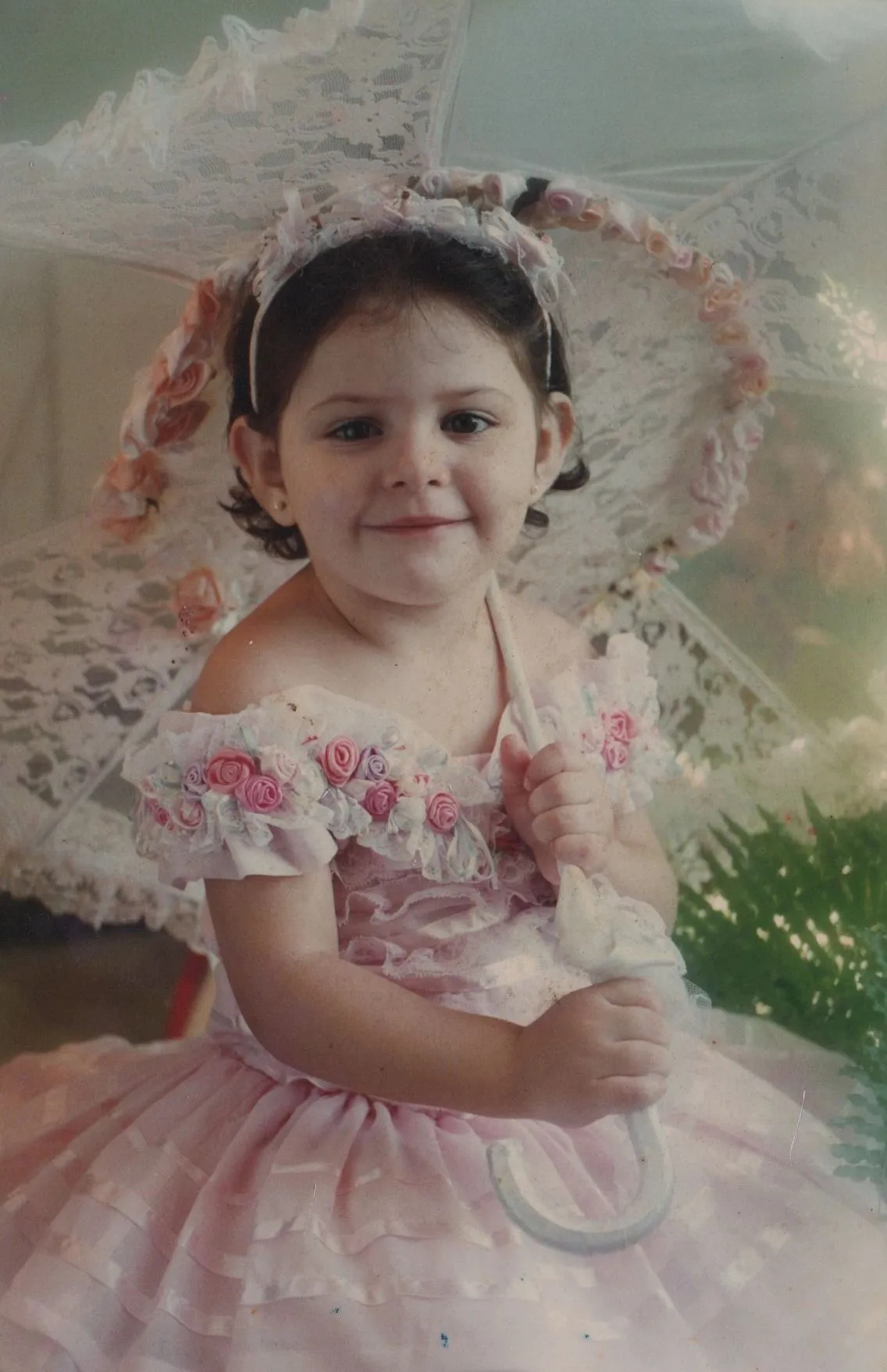 Uma jovem garota com cabelos escuros e um sorriso no rosto, vestindo um vestido de renda rosa pálido e segurando um chapéu de sol de renda branco, sentada contra um fundo de folhagem verde. (Original)