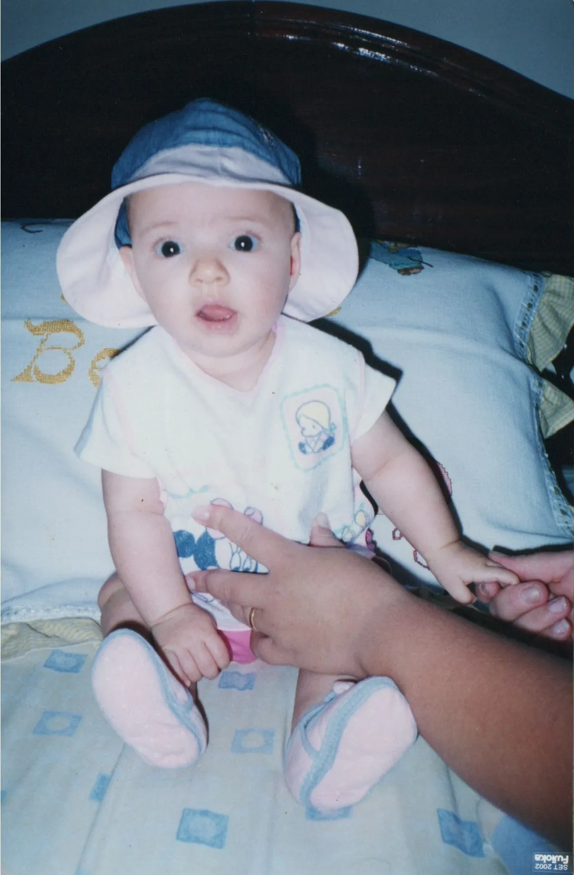 Um bebê de olhos arregalados, com um chapéu e uma camiseta rosa, está sentado com a boca ligeiramente aberta enquanto uma mão adulta segura seus pés. (Original)