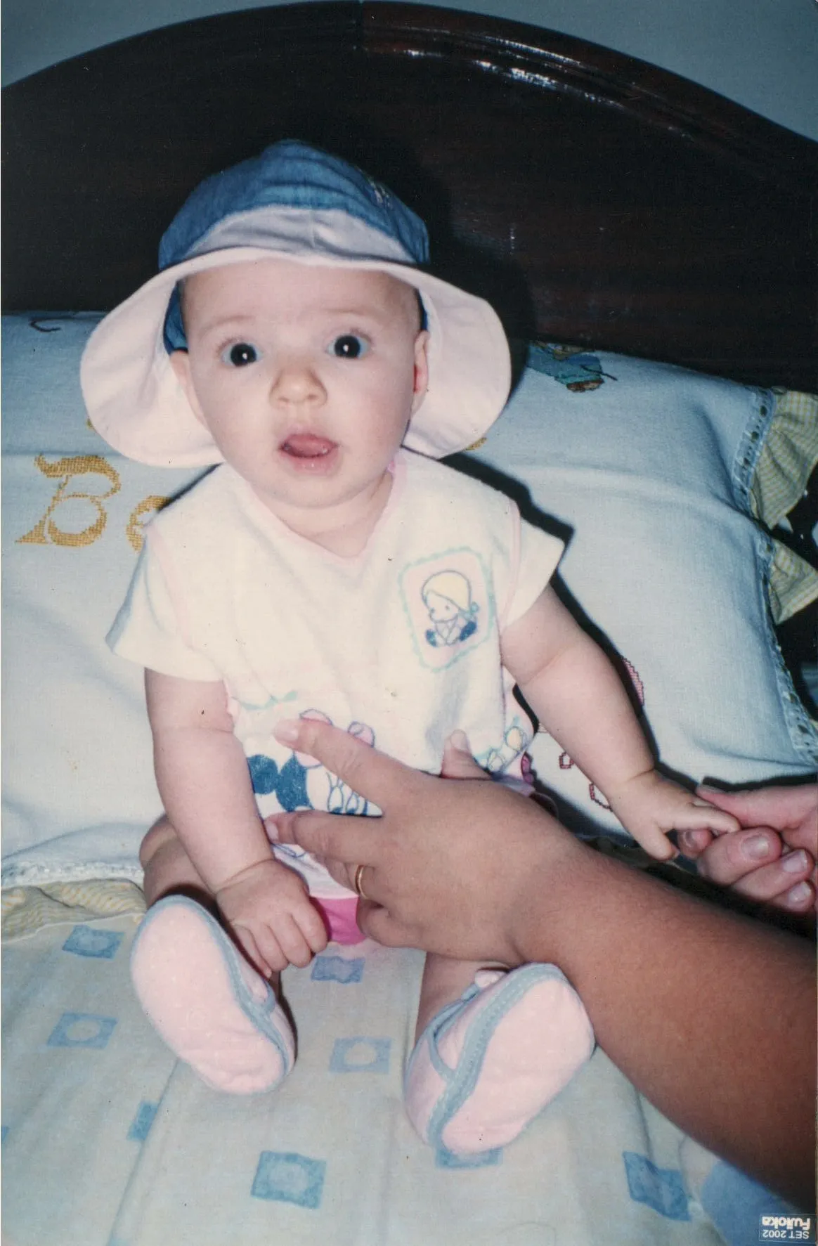 Um bebê de olhos arregalados, com um chapéu e uma camiseta rosa, está sentado com a boca ligeiramente aberta enquanto uma mão adulta segura seus pés.