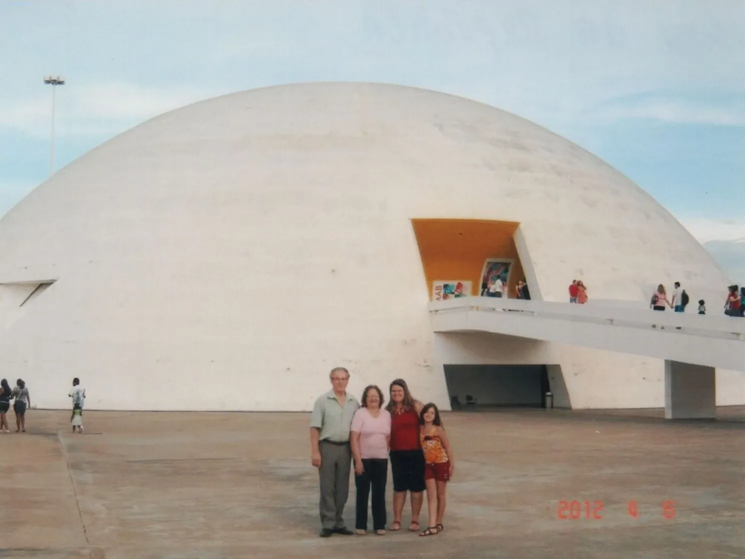 Uma família de quatro pessoas, um homem e três mulheres de diferentes gerações, posa na frente de um edifício branco em forma de cúpula com uma entrada amarela e um caminho elevado.