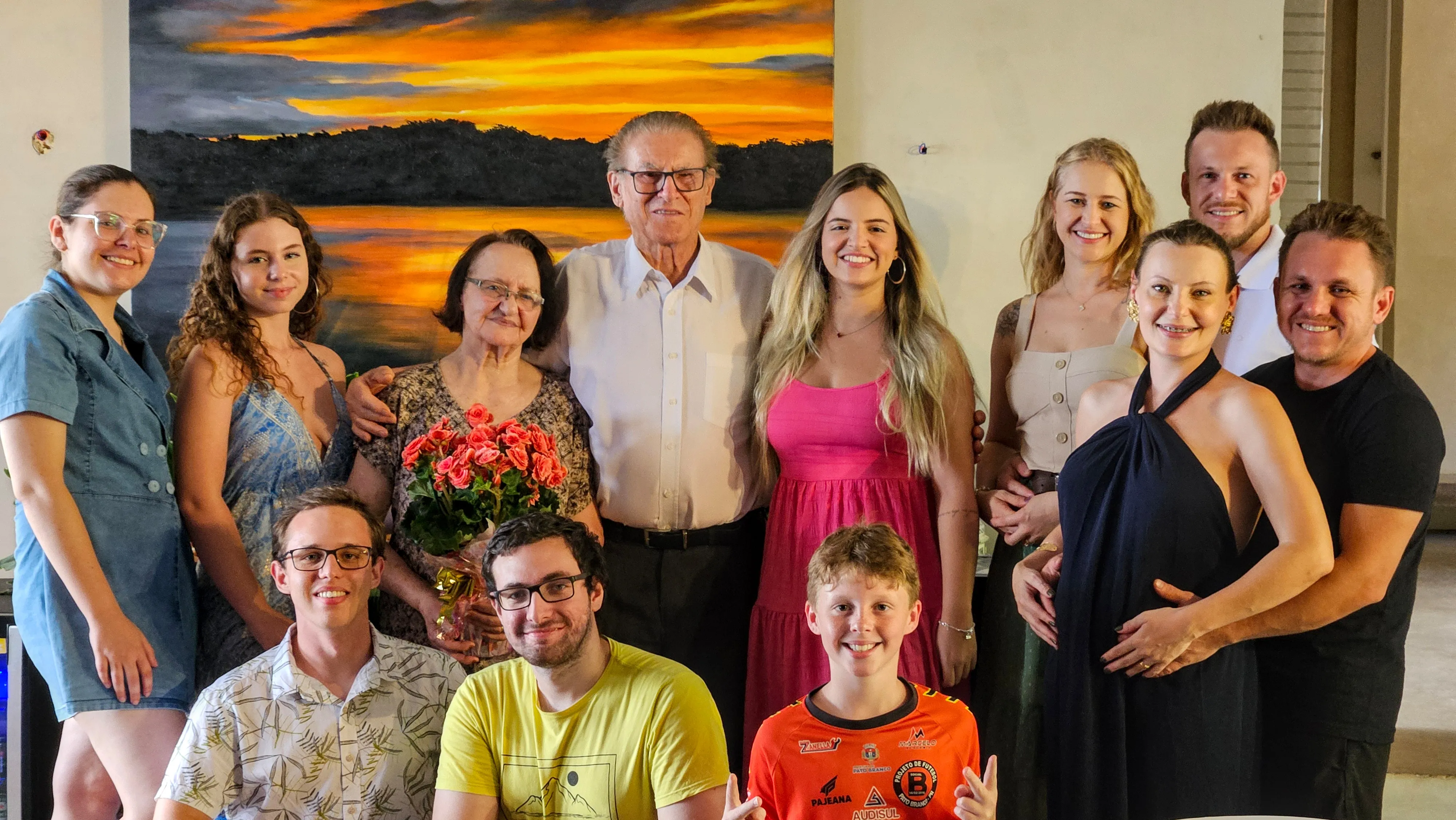 Treze pessoas sorrindo para a câmera em uma foto de família, com uma grande pintura do pôr do sol na parede atrás.