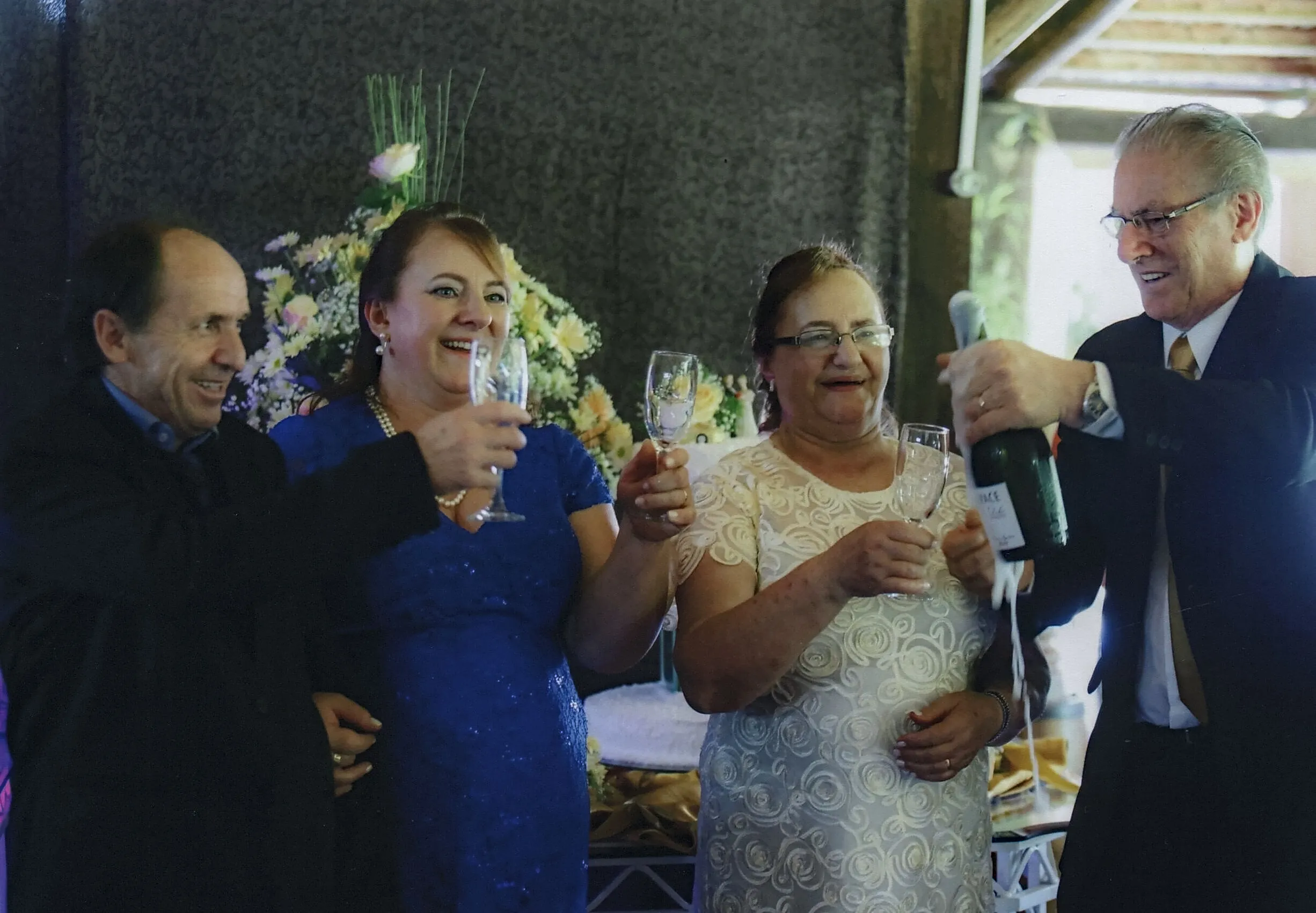 Quatro pessoas brindam com taças de champanhe; um homem de óculos abre uma garrafa de champanhe enquanto os outros três sorriem.
