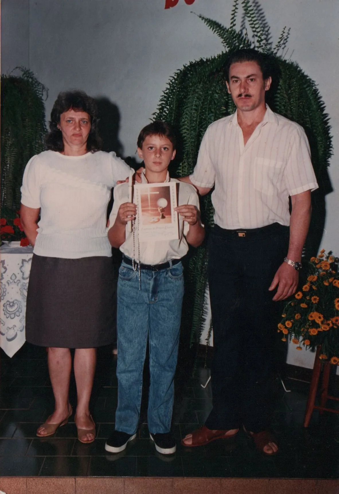 Um homem, uma mulher e uma criança em pé, o homem e a mulher de cada lado da criança, todos olhando para a frente. A criança está segurando um documento. (Original)