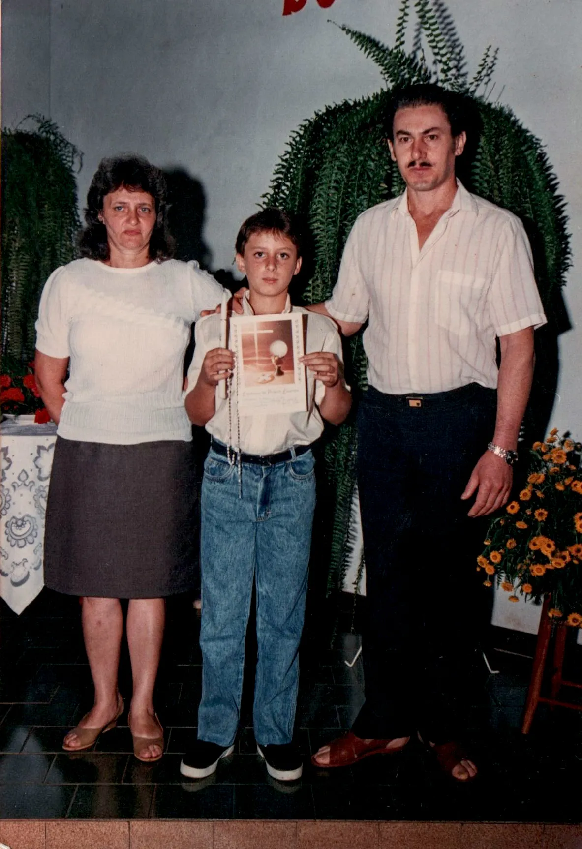 Um homem, uma mulher e uma criança em pé, o homem e a mulher de cada lado da criança, todos olhando para a frente. A criança está segurando um documento.