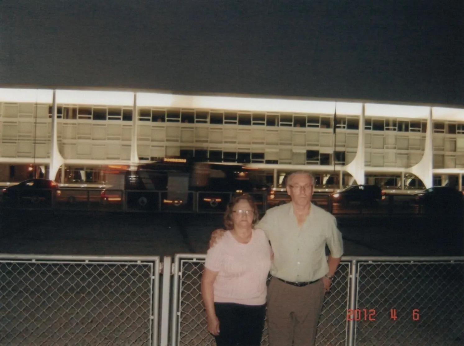 Um casal idoso, com a mulher à esquerda e o homem à direita, posa em frente a um edifício moderno e iluminado à noite.