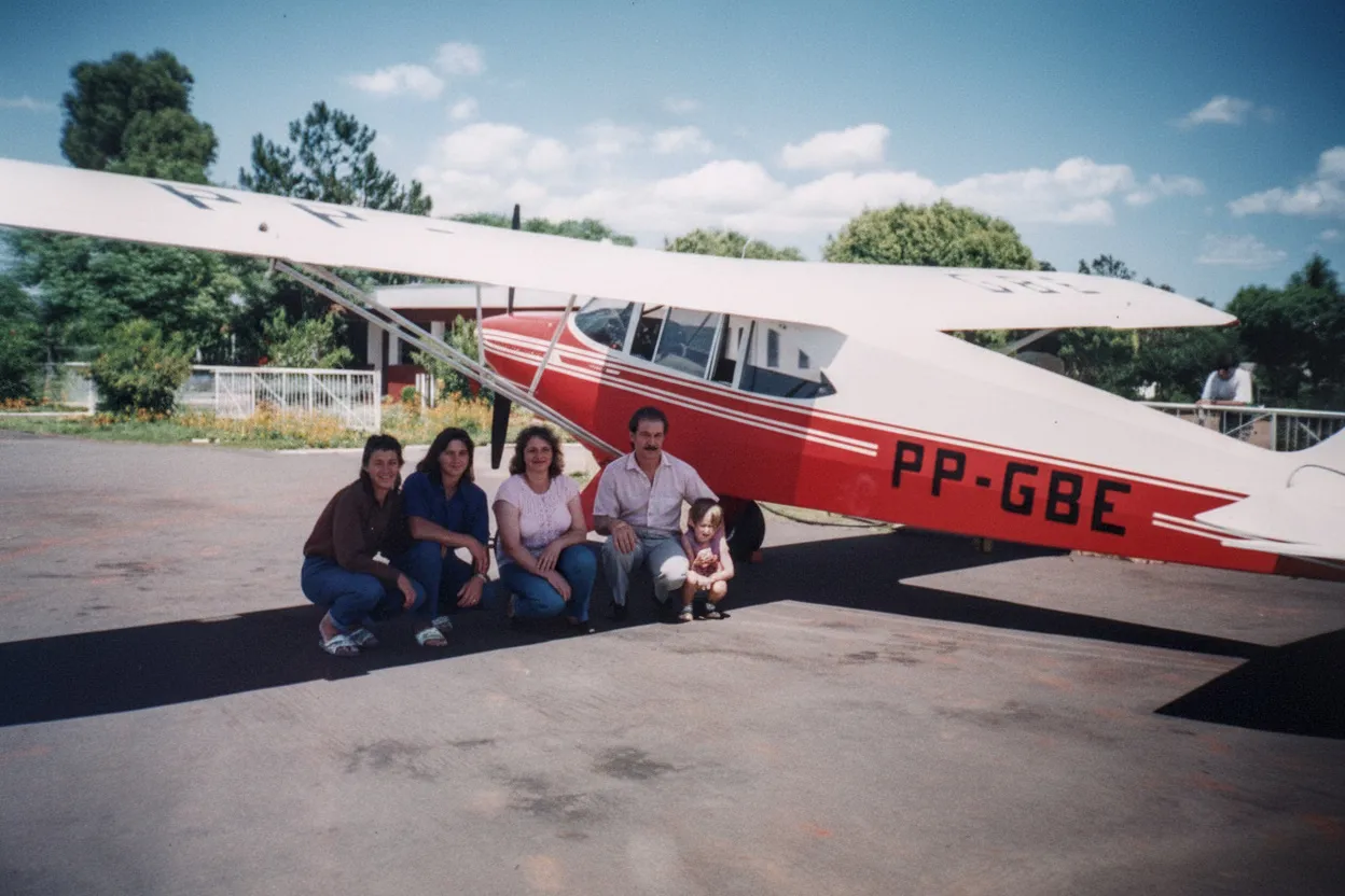Grupo de cinco pessoas, sendo quatro adultos e uma criança, agachadas em frente a um avião monomotor vermelho e branco estacionado ao ar livre.