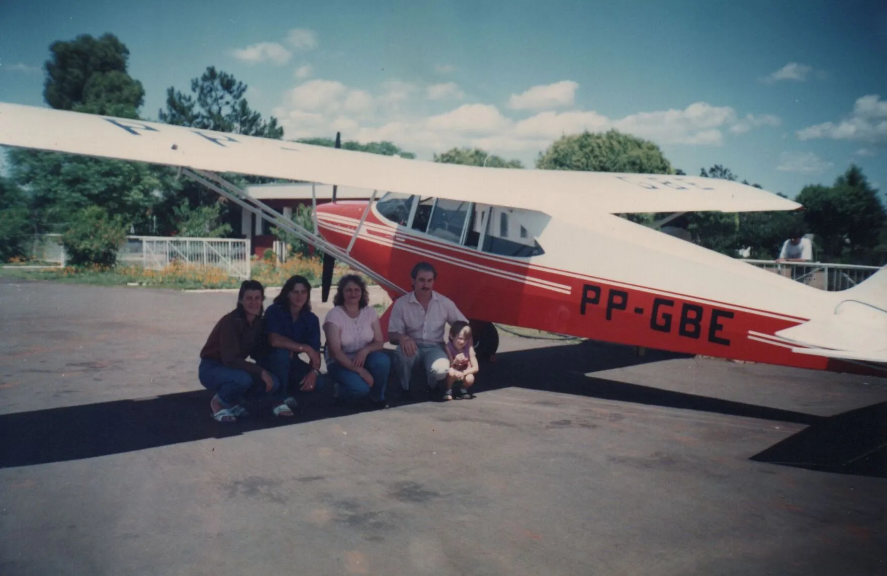 Grupo de cinco pessoas, sendo quatro adultos e uma criança, agachadas em frente a um avião monomotor vermelho e branco estacionado ao ar livre. (Original)
