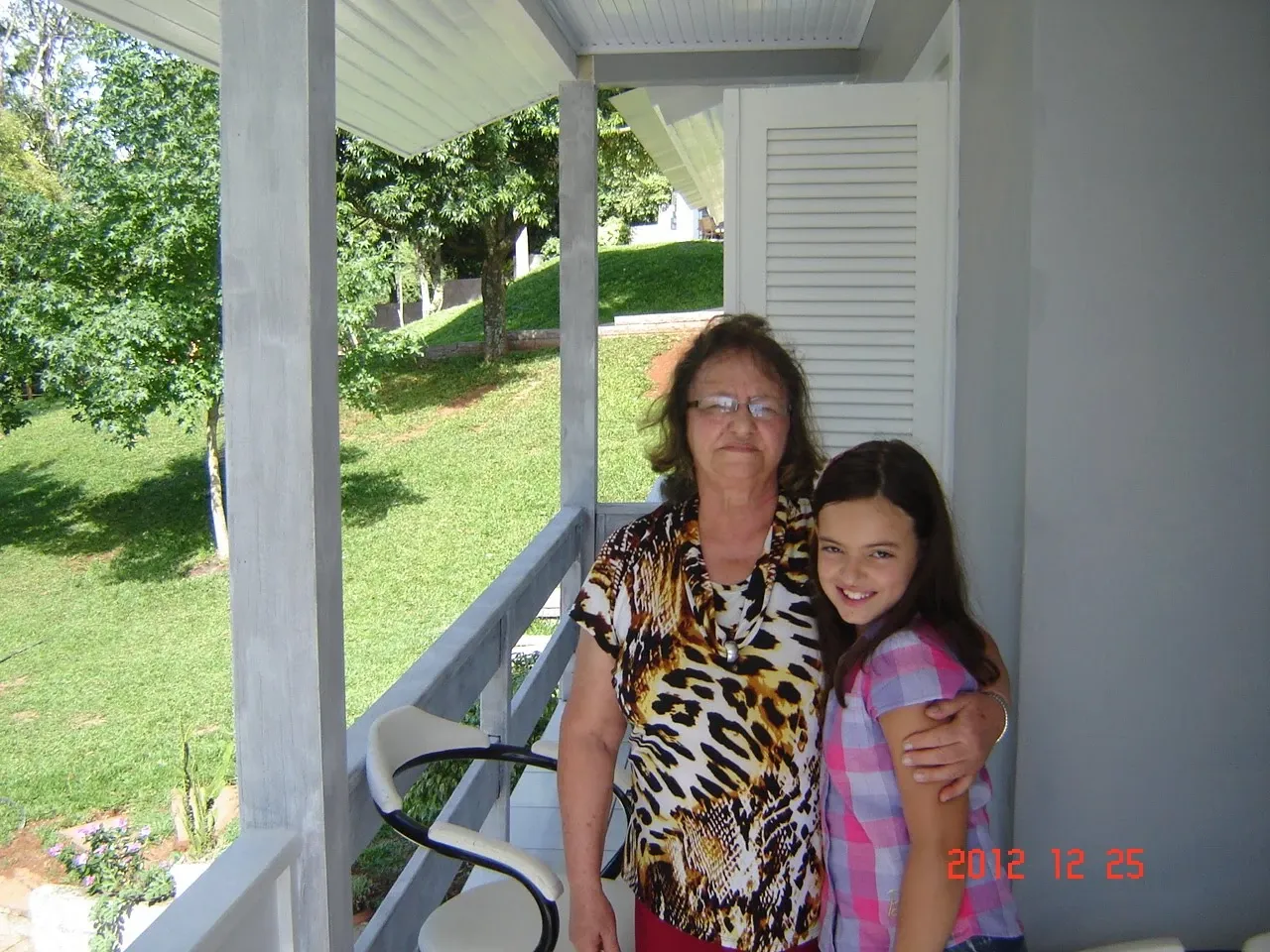 Uma avó e sua neta abraçadas, sorrindo para a câmera em uma varanda iluminada pelo sol, com um gramado verde e árvores ao fundo.
