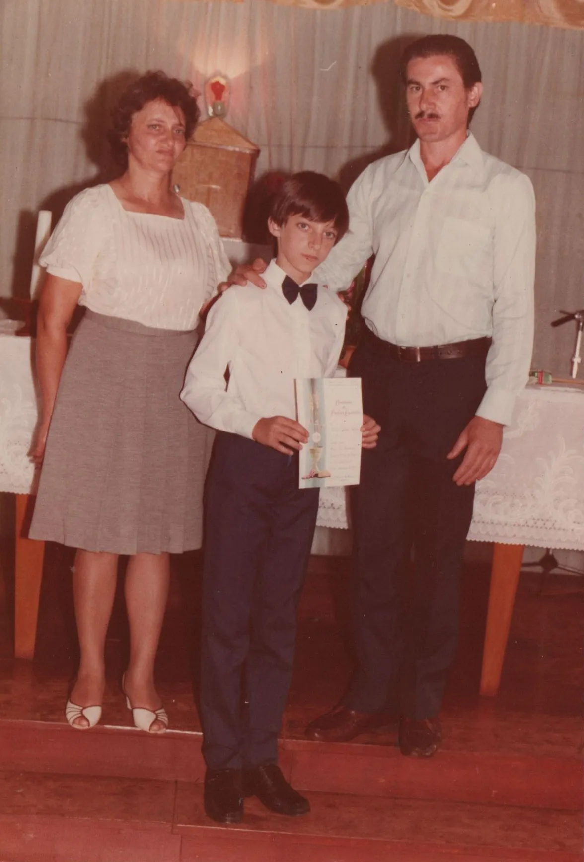 Um homem e uma mulher em roupas formais de pé com um menino em um smoking com uma gravata borboleta e um certificado. (Original)