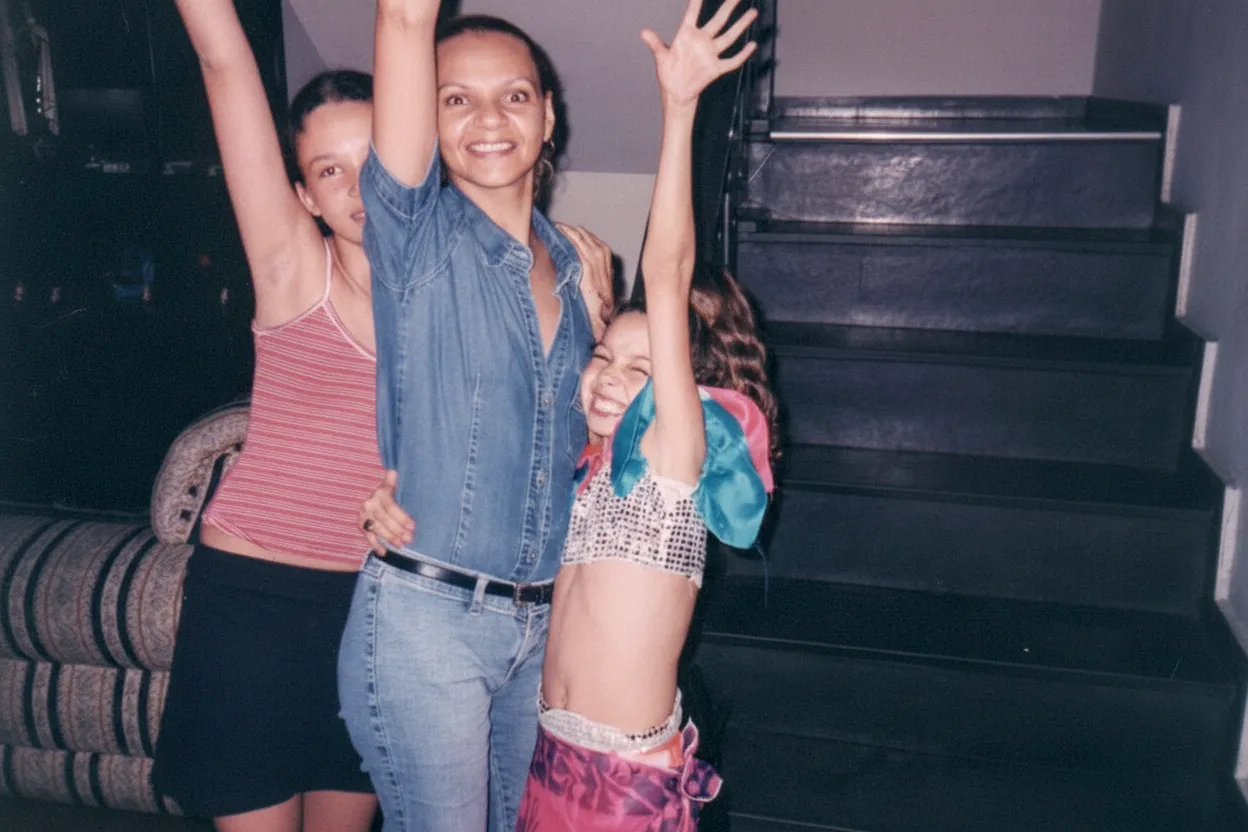 Três mulheres, uma adulta e duas crianças, posam para uma foto em um quarto, sorrindo e levantando os braços.