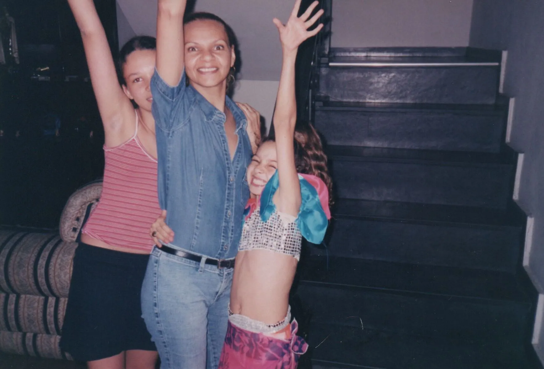 Três mulheres, uma adulta e duas crianças, posam para uma foto em um quarto, sorrindo e levantando os braços. (Original)