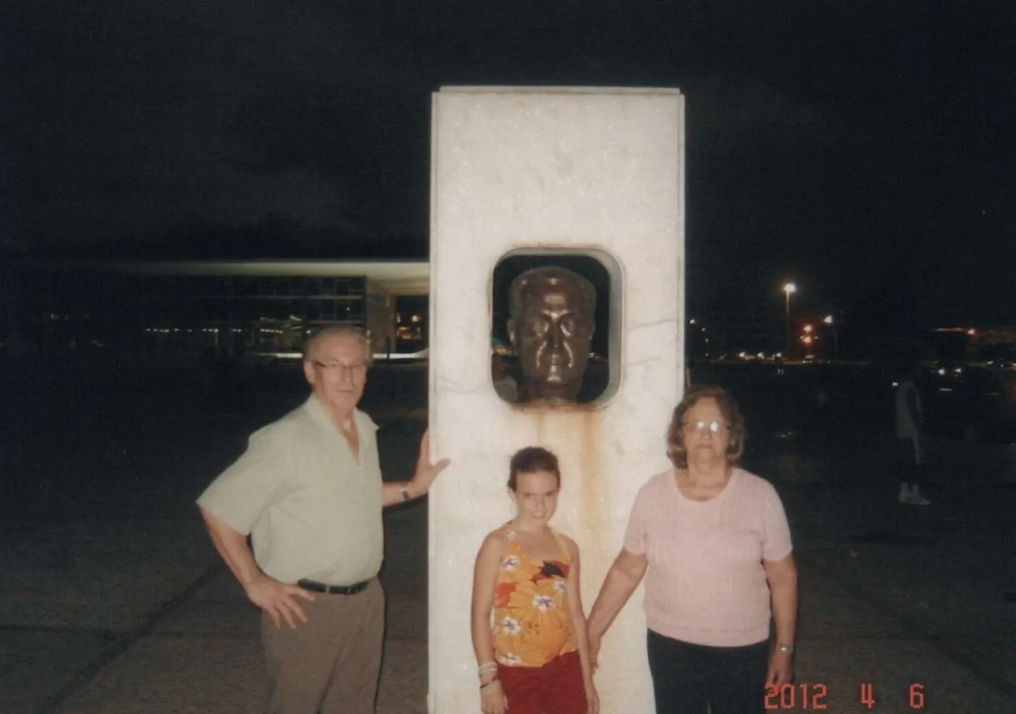 Um homem, uma mulher e uma menina estão parados ao lado de um busto de bronze dentro de uma estrutura retangular branca, à noite.