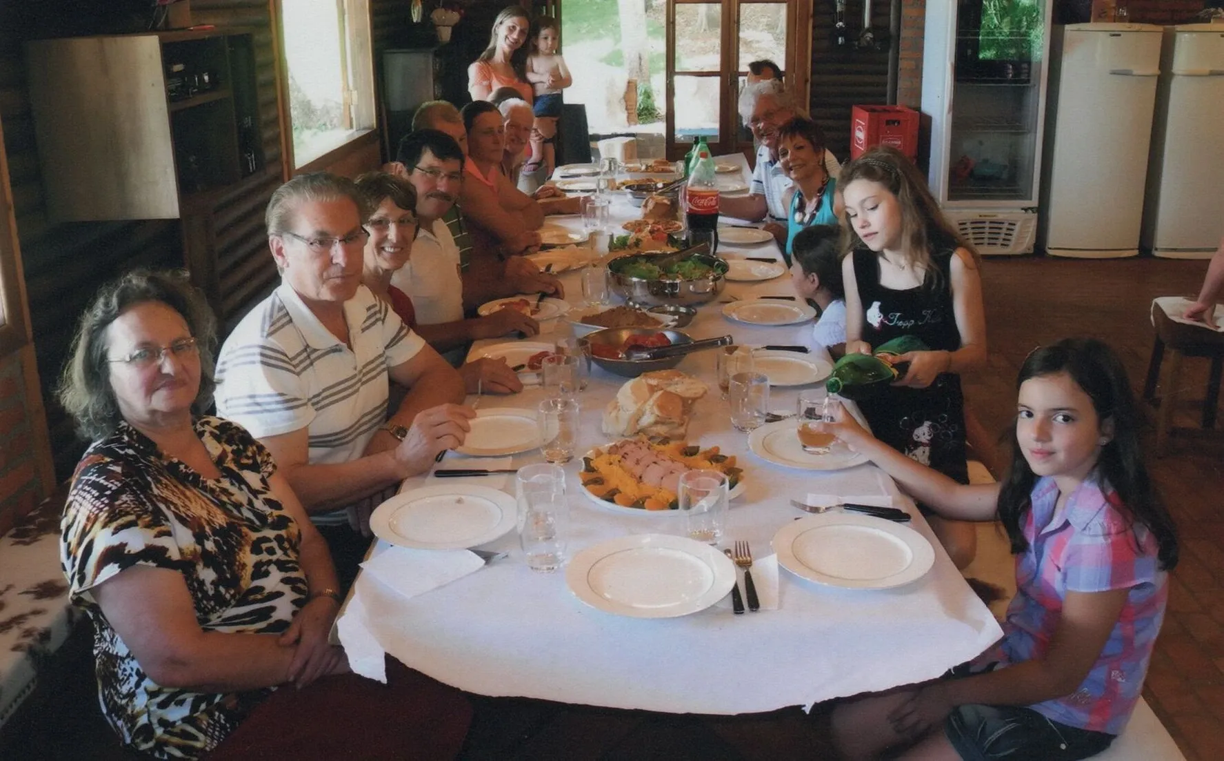 Uma família numerosa reunida em torno de uma mesa branca para uma refeição informal.