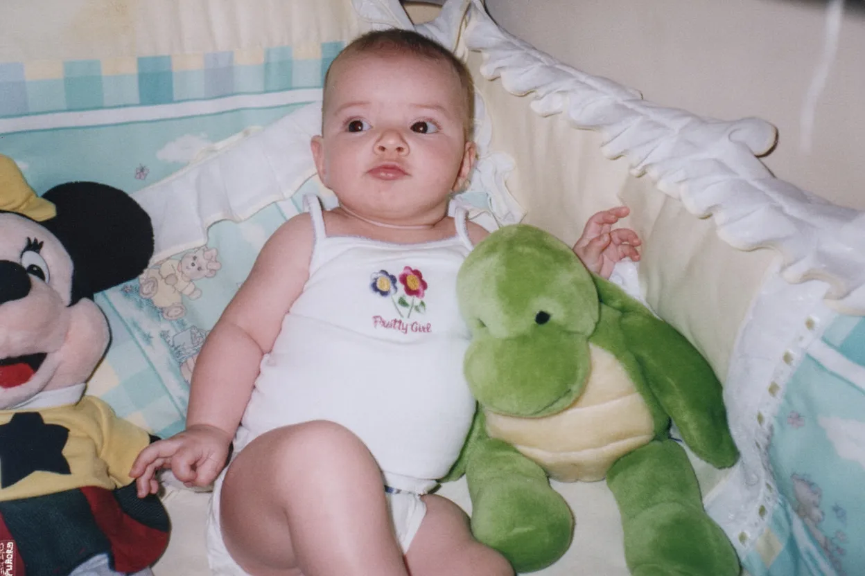 Um bebê de pele clara, deitado de costas em um berço, olhando para cima com olhos arregalados. Ele está vestindo um body branco e um fralda, com um bicho de pelúcia do Mickey Mouse à sua esquerda e uma tartaruga verde à sua direita.