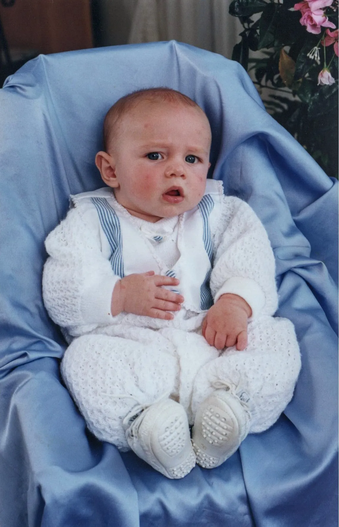 Bebê de pele clara sentado em uma roupa de malha branca com suspensórios azuis, sentado em um cobertor azul com plantas cor-de-rosa ao fundo.