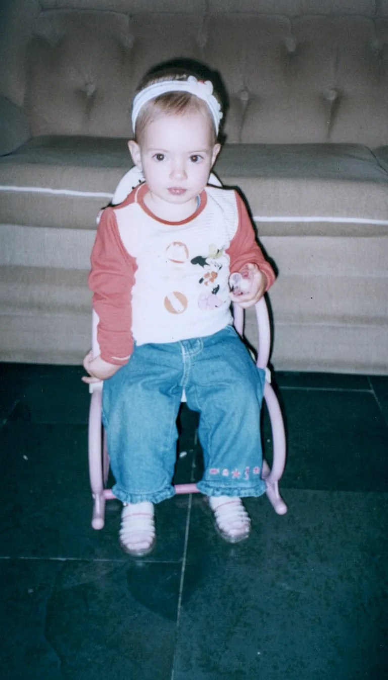 Bebê sentado em uma pequena cadeira rosa em um quarto escuro com um sofá bege ao fundo.