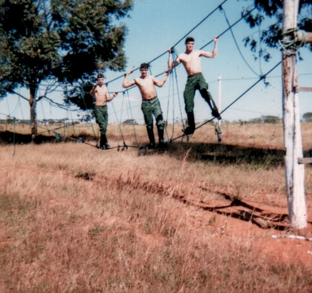 Três soldados sem camisa, usando calças e botas militares, navegam por uma ponte de corda instável sobre um campo gramado e seco com árvores ao fundo.