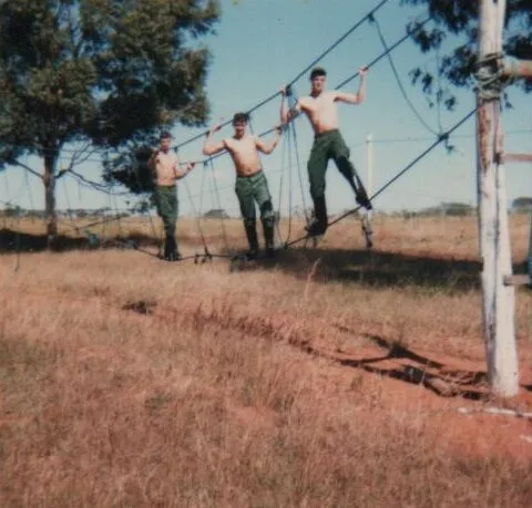 Três soldados sem camisa, usando calças e botas militares, navegam por uma ponte de corda instável sobre um campo gramado e seco com árvores ao fundo. (Original)