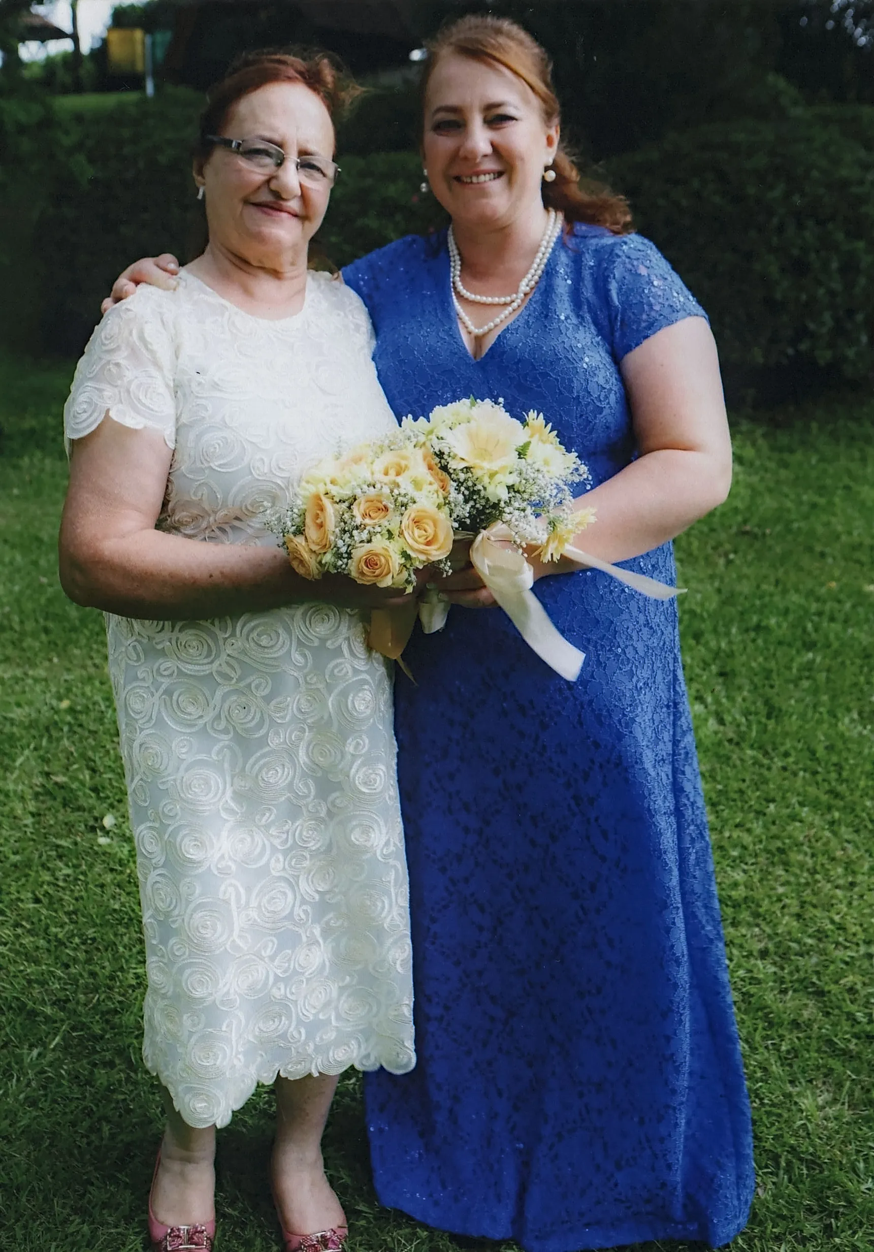 Duas mulheres, uma mais velha de vestido branco e outra mais jovem de vestido azul, estão abraçadas e sorrindo ao ar livre, segurando um buquê de flores.