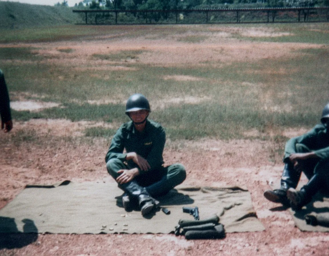 Um soldado vestido com um uniforme verde-oliva e capacete está sentado de pernas cruzadas em uma lona no chão, com um revólver e munição ao seu lado, em um campo de tiro de terra e grama.