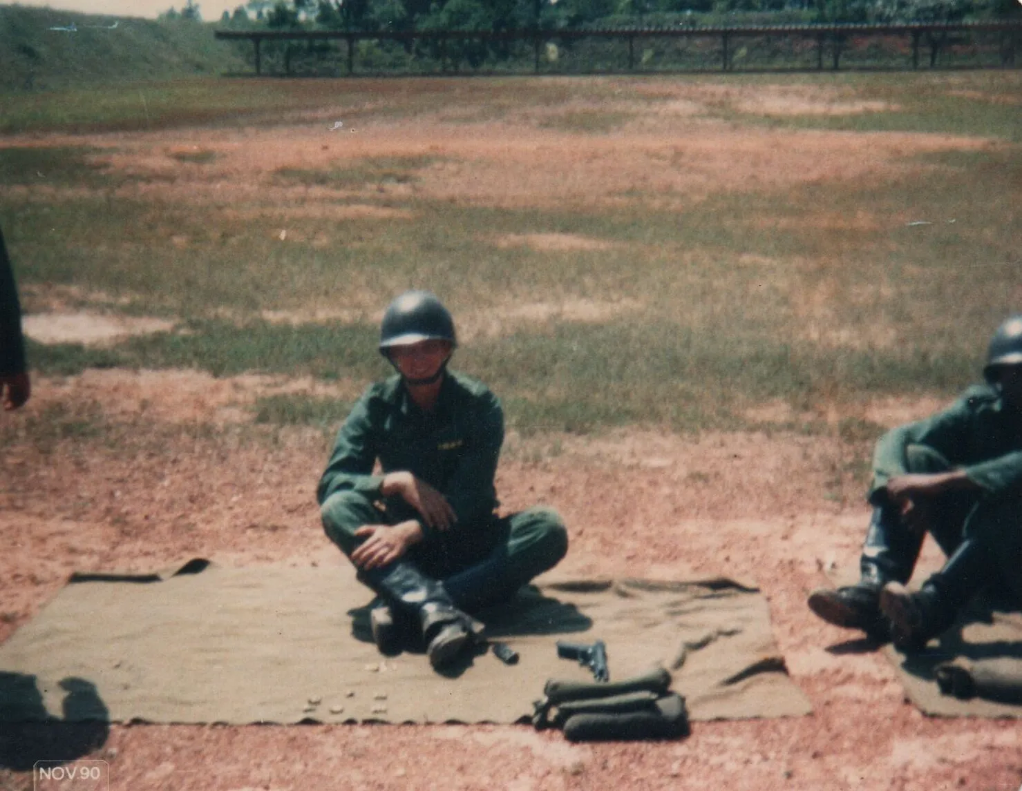 Um soldado vestido com um uniforme verde-oliva e capacete está sentado de pernas cruzadas em uma lona no chão, com um revólver e munição ao seu lado, em um campo de tiro de terra e grama. (Original)