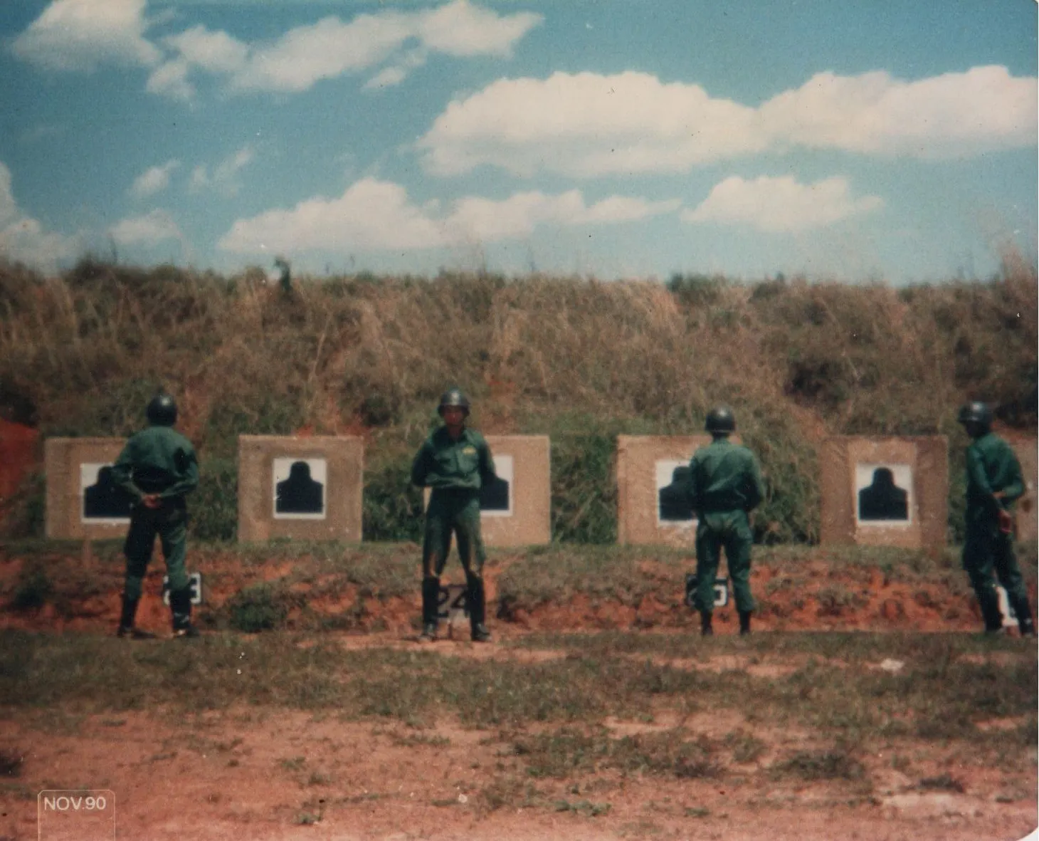 Quatro soldados em uniformes verdes, voltados para alvos de tiro ao ar livre sob um céu azul. (Original)