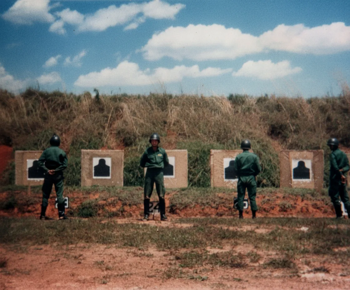 Quatro soldados em uniformes verdes, voltados para alvos de tiro ao ar livre sob um céu azul.