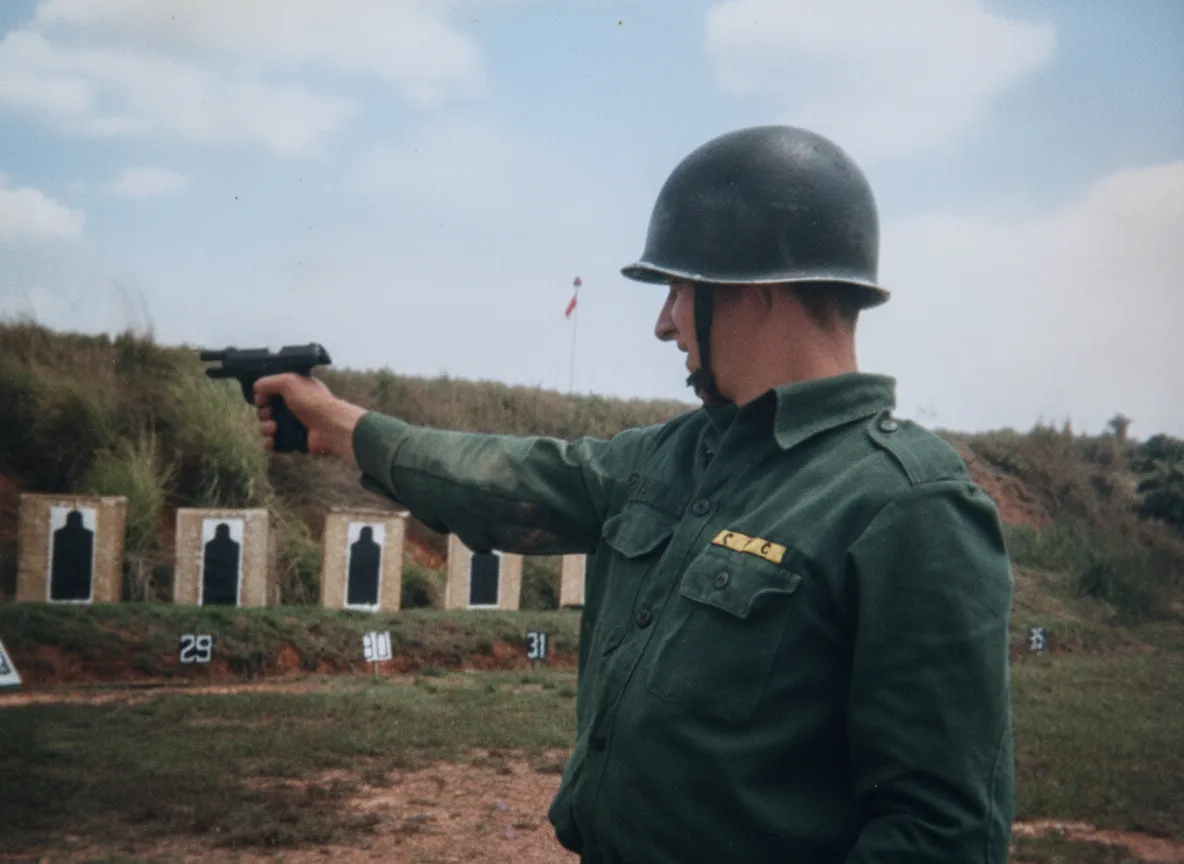 Um soldado com capacete verde-oliva e camisa de manga comprida atira uma pistola preta na lateral de um campo de tiro contra vários alvos de silhueta.