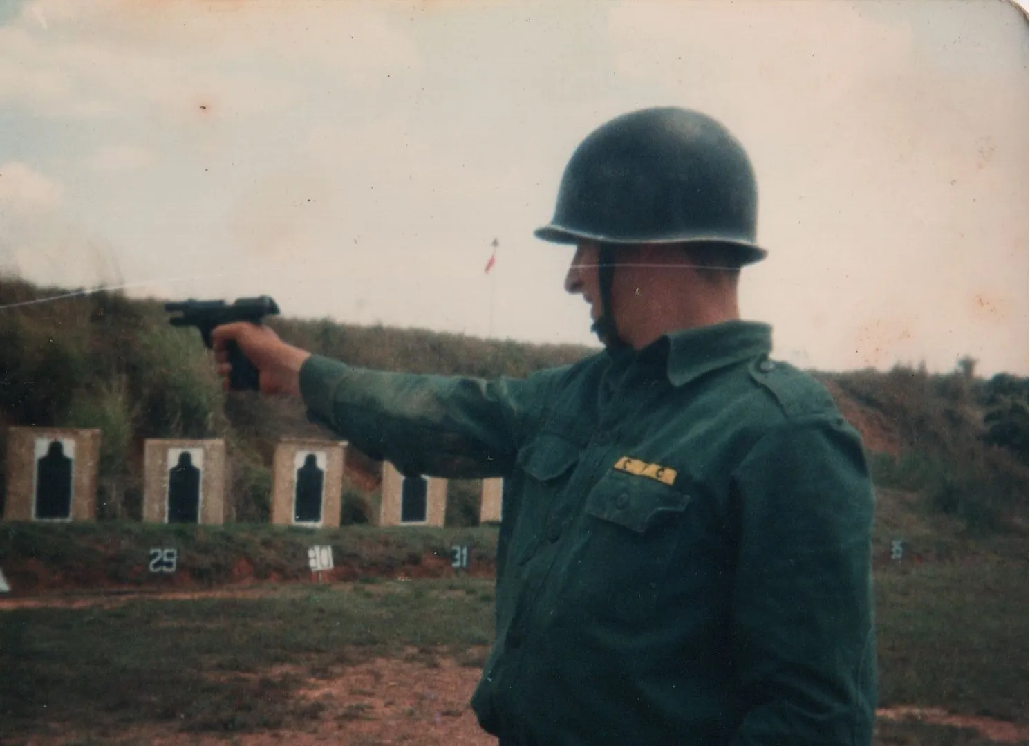 Um soldado com capacete verde-oliva e camisa de manga comprida atira uma pistola preta na lateral de um campo de tiro contra vários alvos de silhueta. (Original)