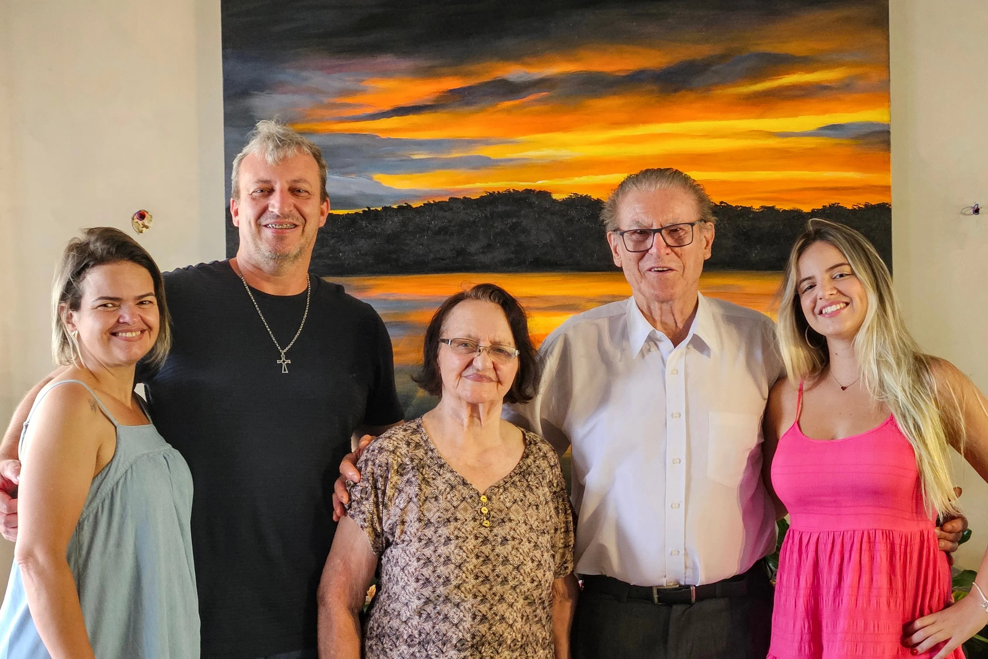 Cinco pessoas, três mulheres e dois homens, com idades variadas, estão em pé em frente a uma grande pintura que retrata um pôr do sol laranja e amarelo sobre uma paisagem escura de montanhas e água.