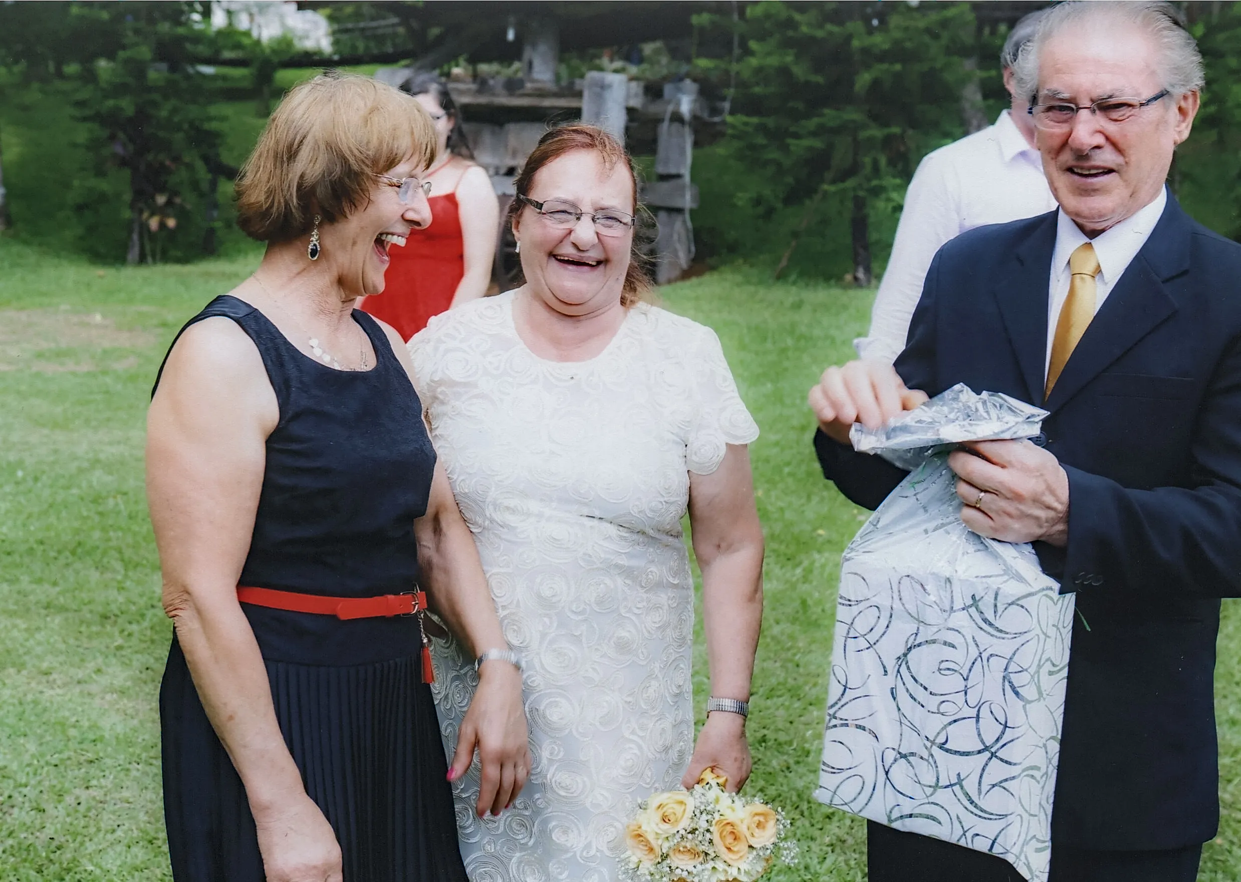 Três adultos sorrindo ao ar livre em um evento: duas mulheres no centro, uma de vestido preto e outra de vestido branco com um buquê, e um homem de terno abrindo um presente à direita.