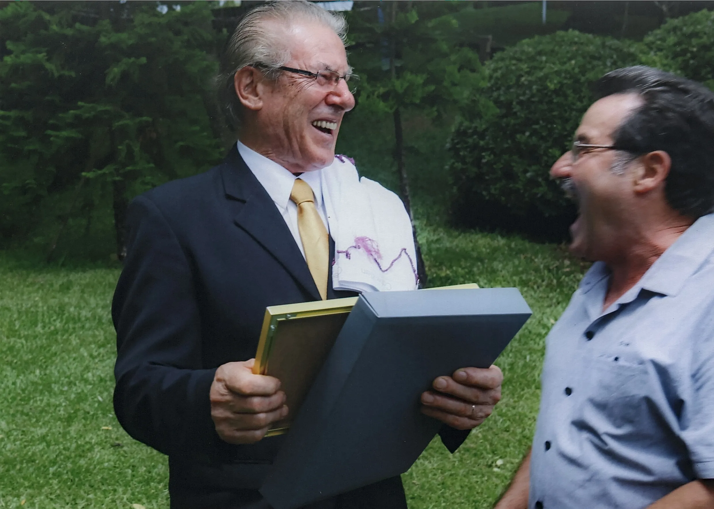 Dois homens em um gramado, o da esquerda em um terno escuro com uma gravata dourada, sorrindo e segurando uma moldura dourada e uma caixa cinza, e o da direita em uma camisa azul-clara, com a boca aberta em uma risada.
