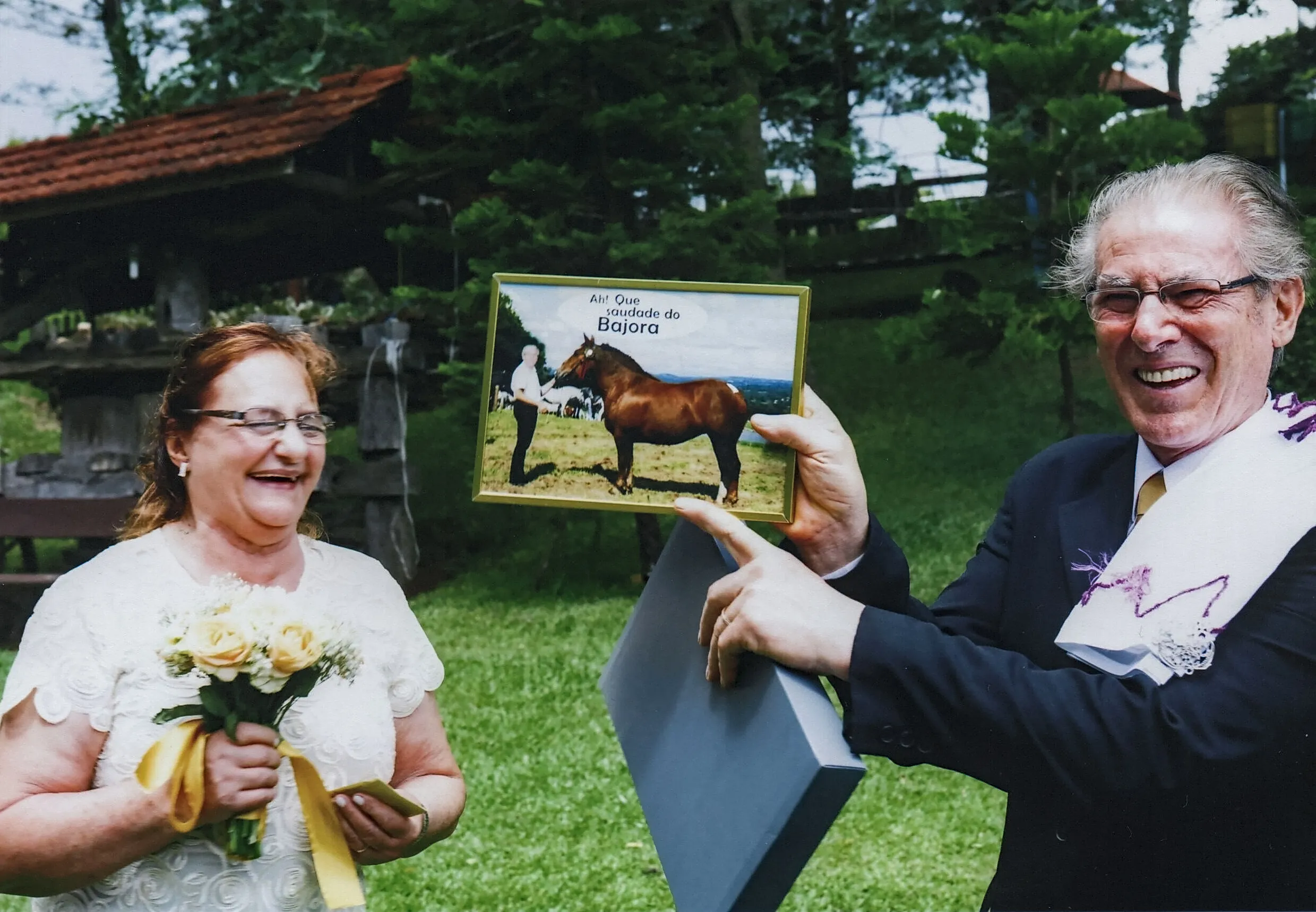 Um homem idoso de terno e óculos segura um porta-retratos com a imagem de um cavalo e um homem, rindo ao lado de uma mulher de vestido branco com um buquê amarelo, em um cenário externo gramado.