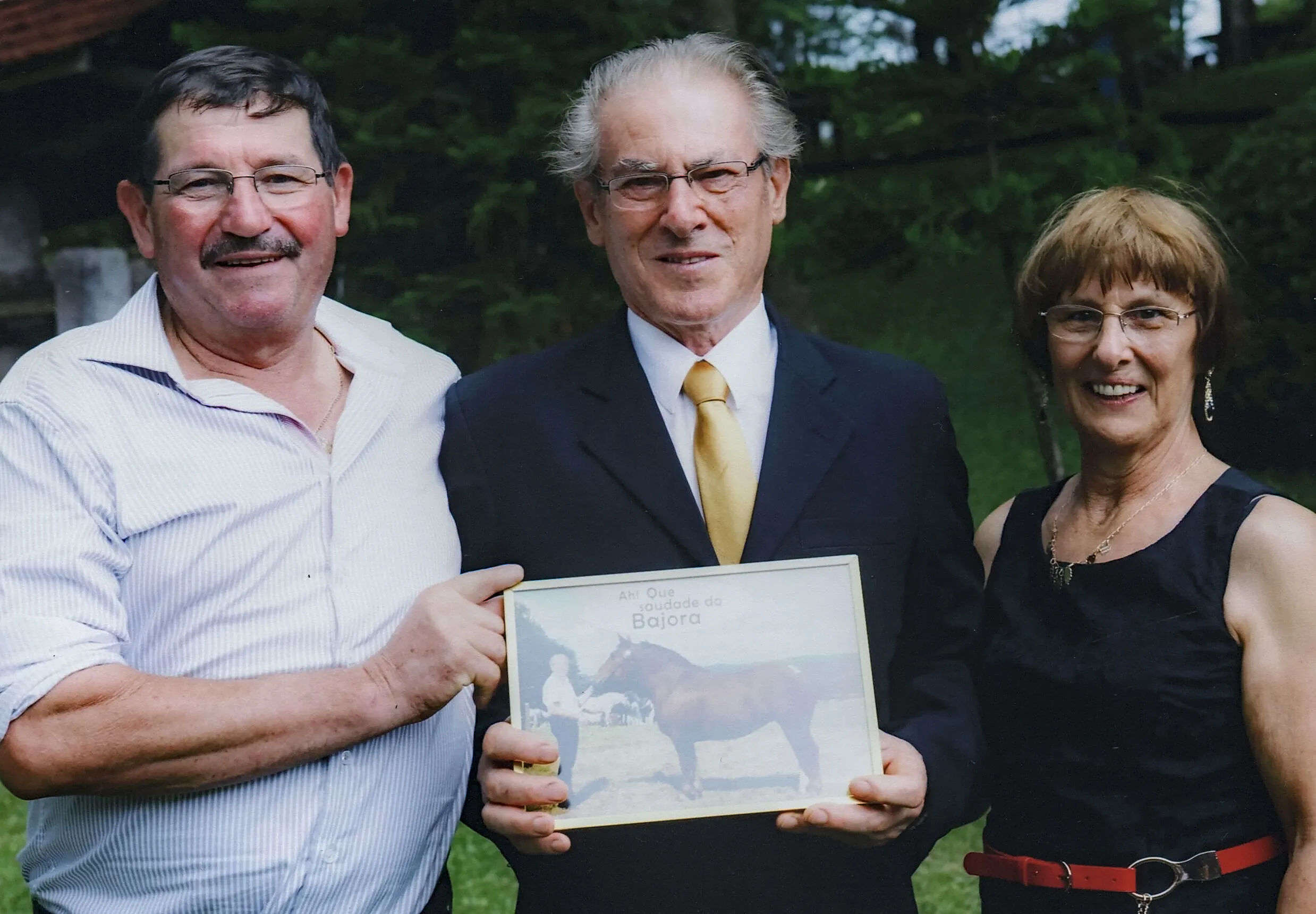 Três pessoas, dois homens e uma mulher, seguram um retrato emoldurado de um cavalo.