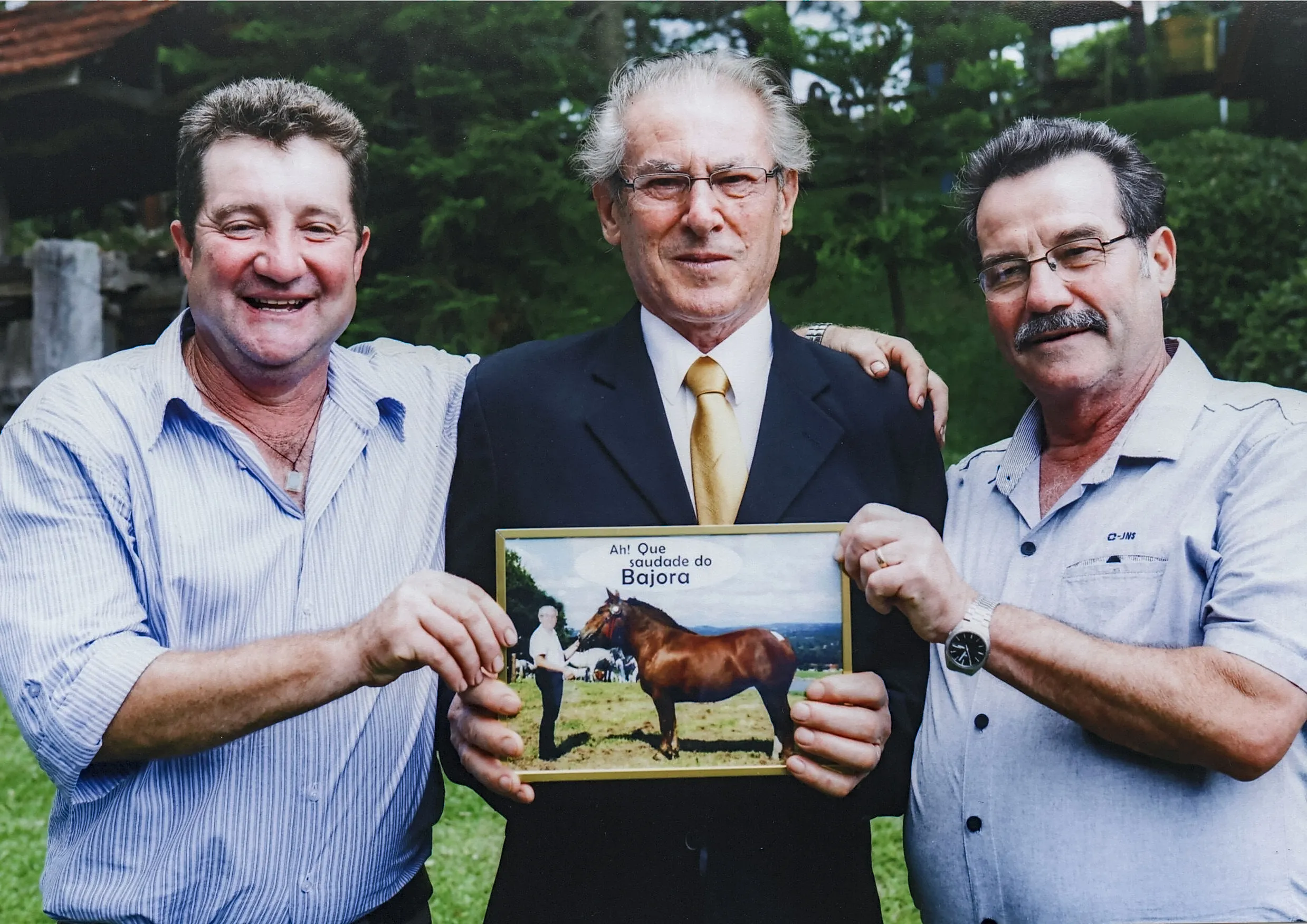 Três homens de meia-idade posam com um quadro de um cavalo no campo; o homem da esquerda sorri para o espectador, o homem do centro sorri para a frente, e o homem da direita tem um bigode e olha para a direita.