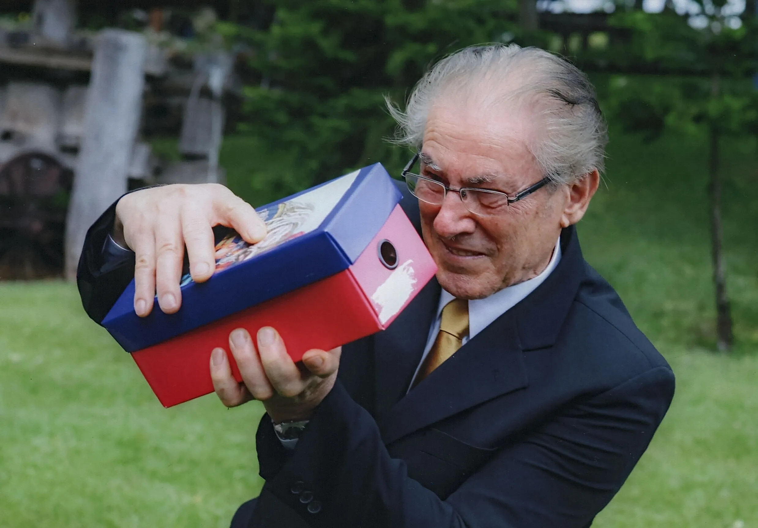 Um homem idoso de óculos e terno escuro tenta equilibrar duas caixas de arquivo coloridas, uma azul e outra vermelha, com uma expressão de esforço.