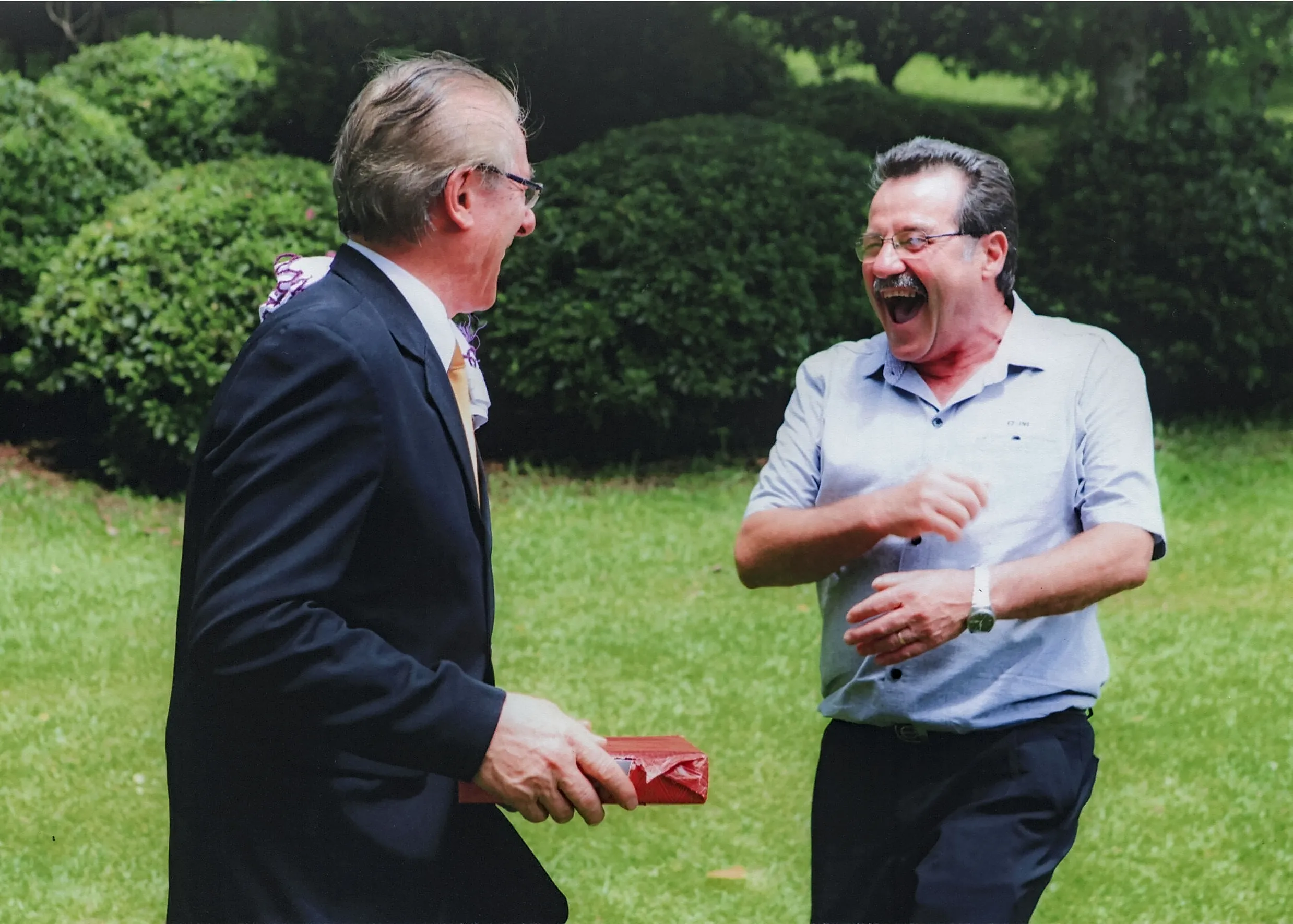 Dois homens sorridentes ao ar livre: um homem de terno segura uma caixa vermelha e se vira para o outro homem, que usa camisa social e está rindo com as mãos na frente do corpo.