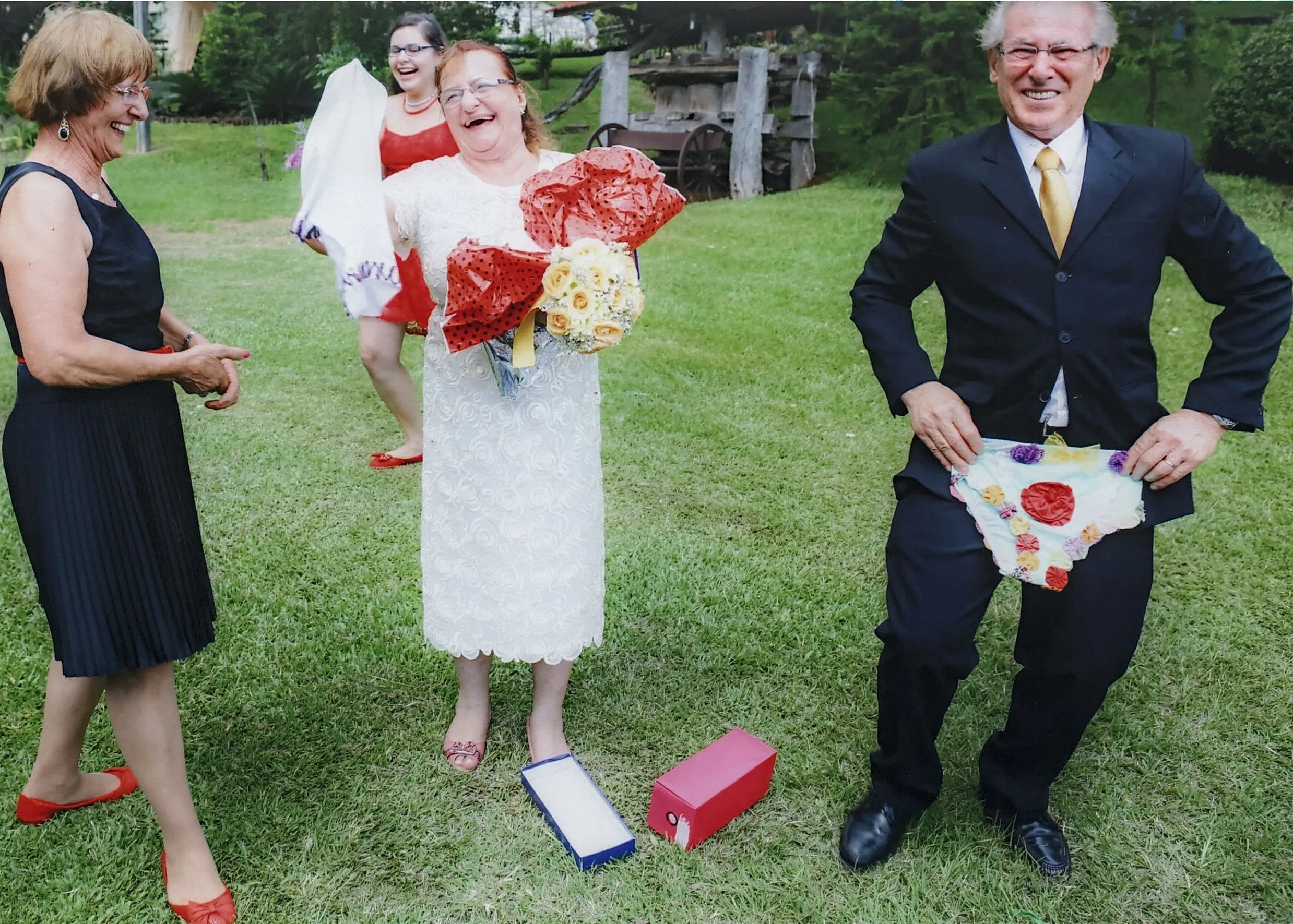 Quatro pessoas sorridentes em um gramado verde, com uma mulher de vestido branco no centro segurando dois buquês e um homem à direita erguendo uma calcinha de bolinhas.