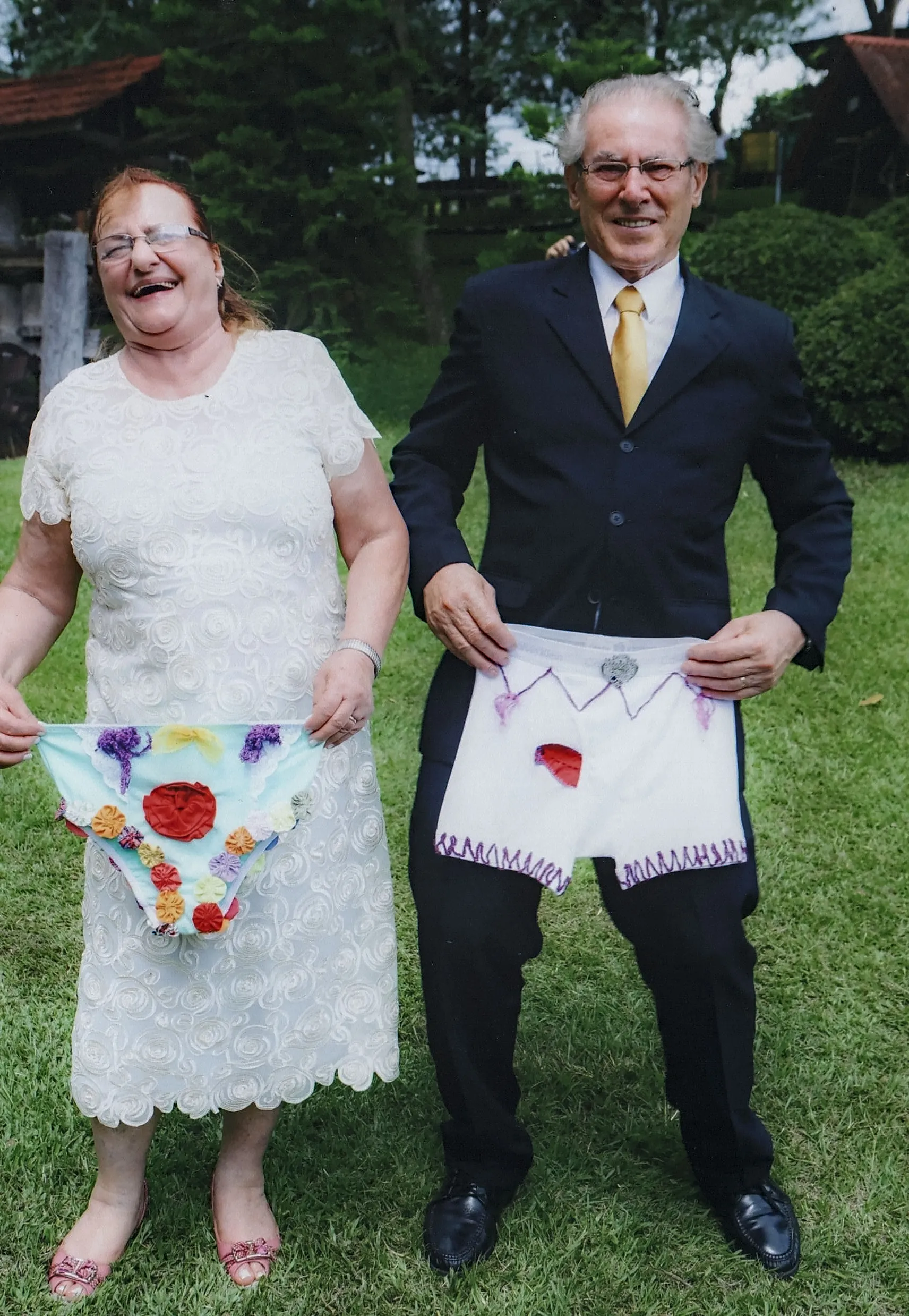Um casal sorridente em um jardim, a mulher em um vestido branco exibindo uma calcinha floral colorida e o homem em um terno preto com gravata amarela segurando uma cueca boxer branca com detalhes roxos e uma mancha vermelha.